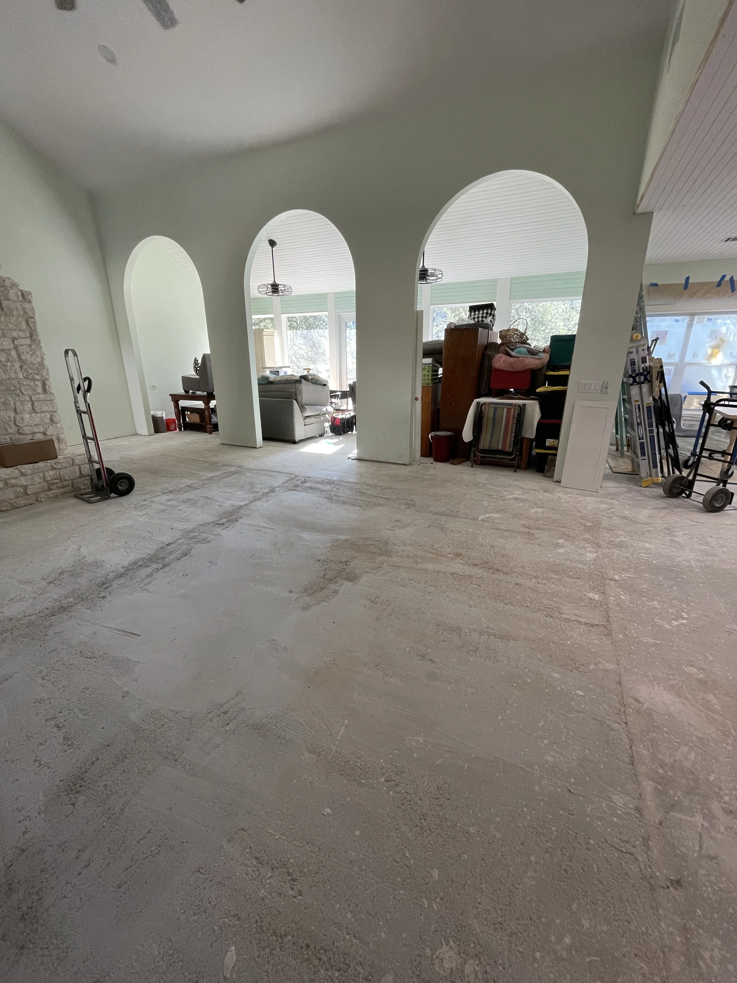 Living room under renovation with unfinished floor, arched doorways, and furniture in the background.