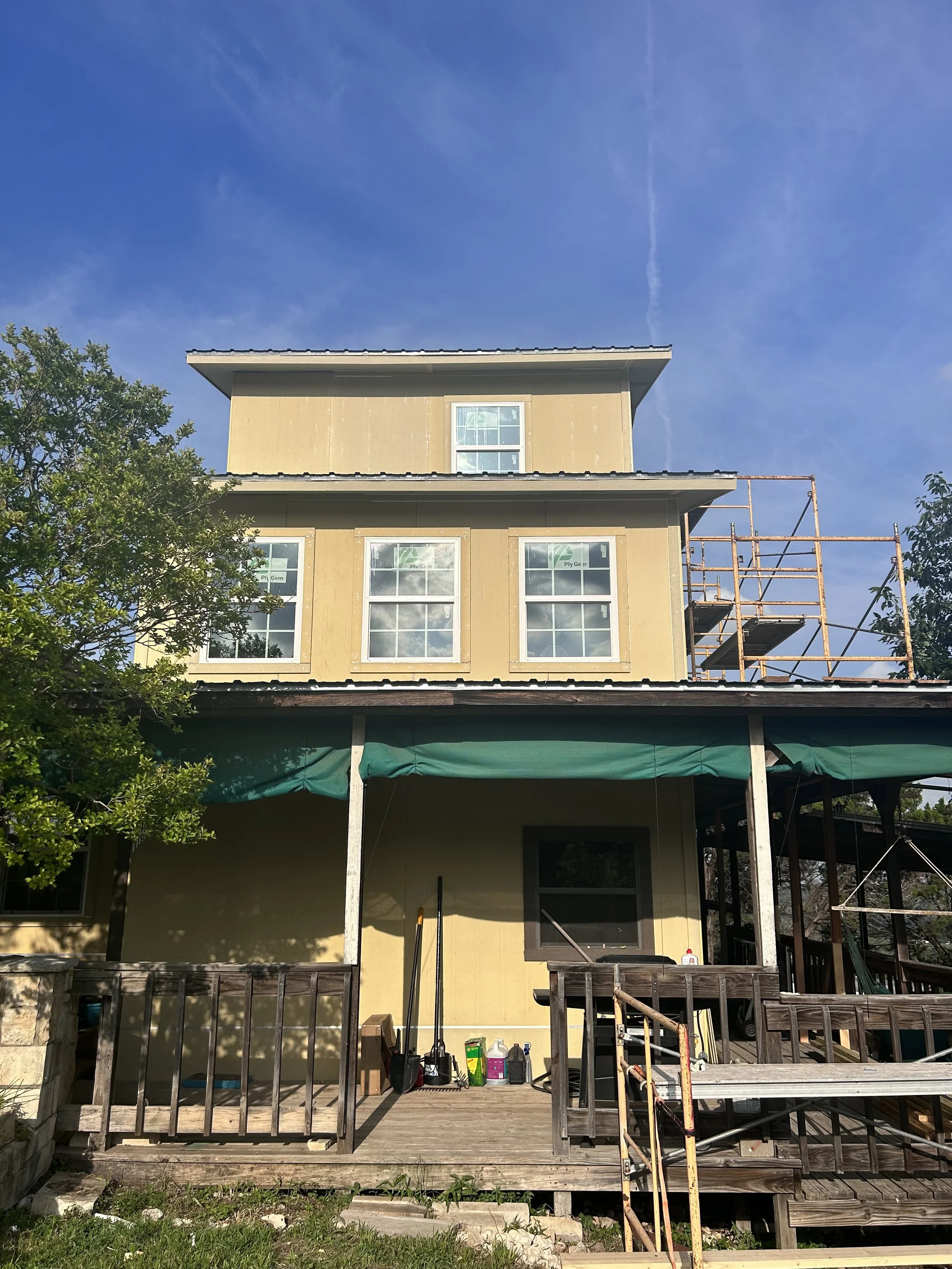 Exterior of a multi-story house under renovation, with scaffolding on the side, a wooden deck, and construction tools and supplies on the deck, under a clear blue sky.