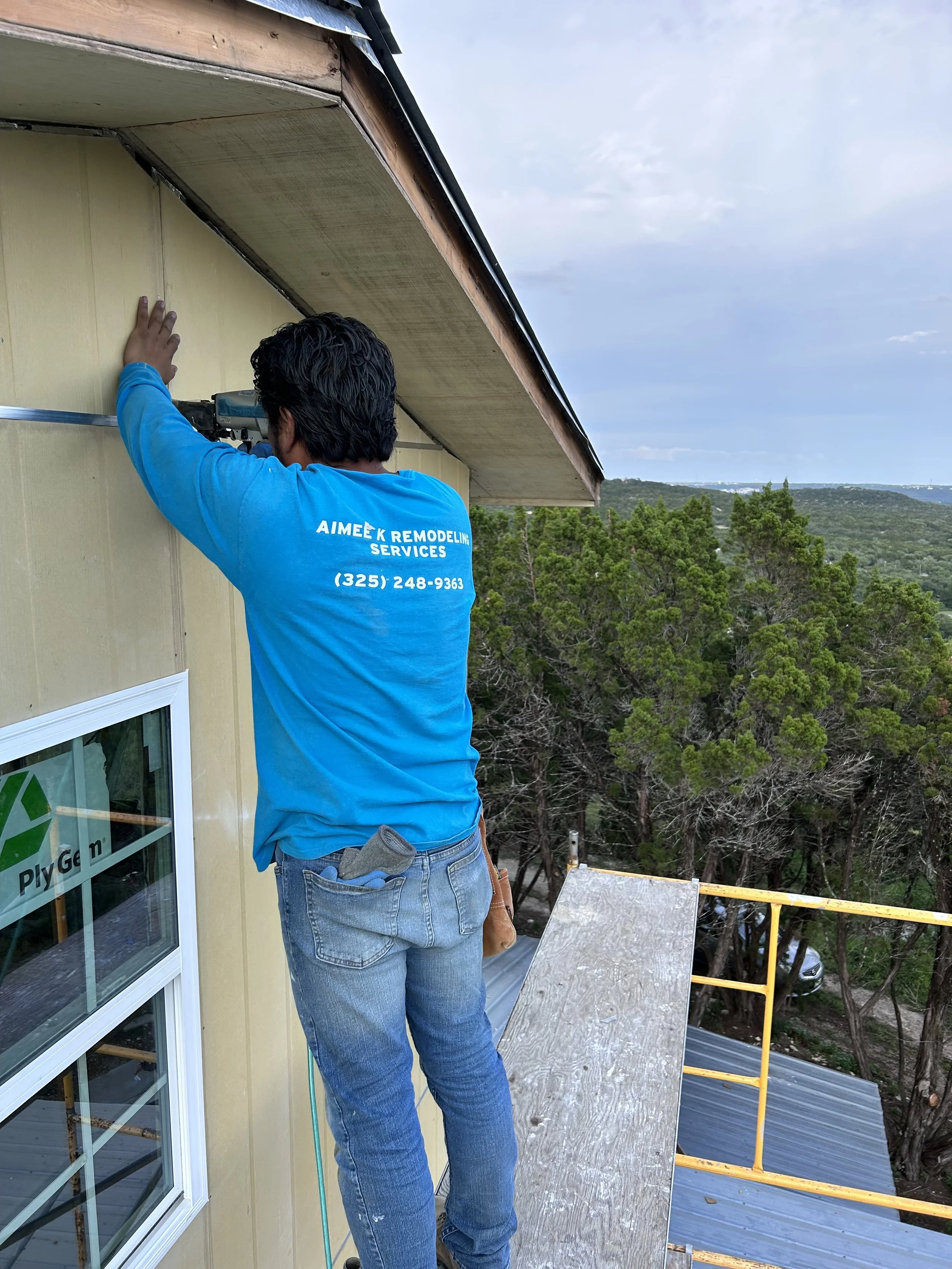 A man wearing a blue shirt with the text 'AIMÉK REMODELING SERVICES' and a phone number on the back, is working on the exterior of a house, fixing or installing something near the roof with a power tool.