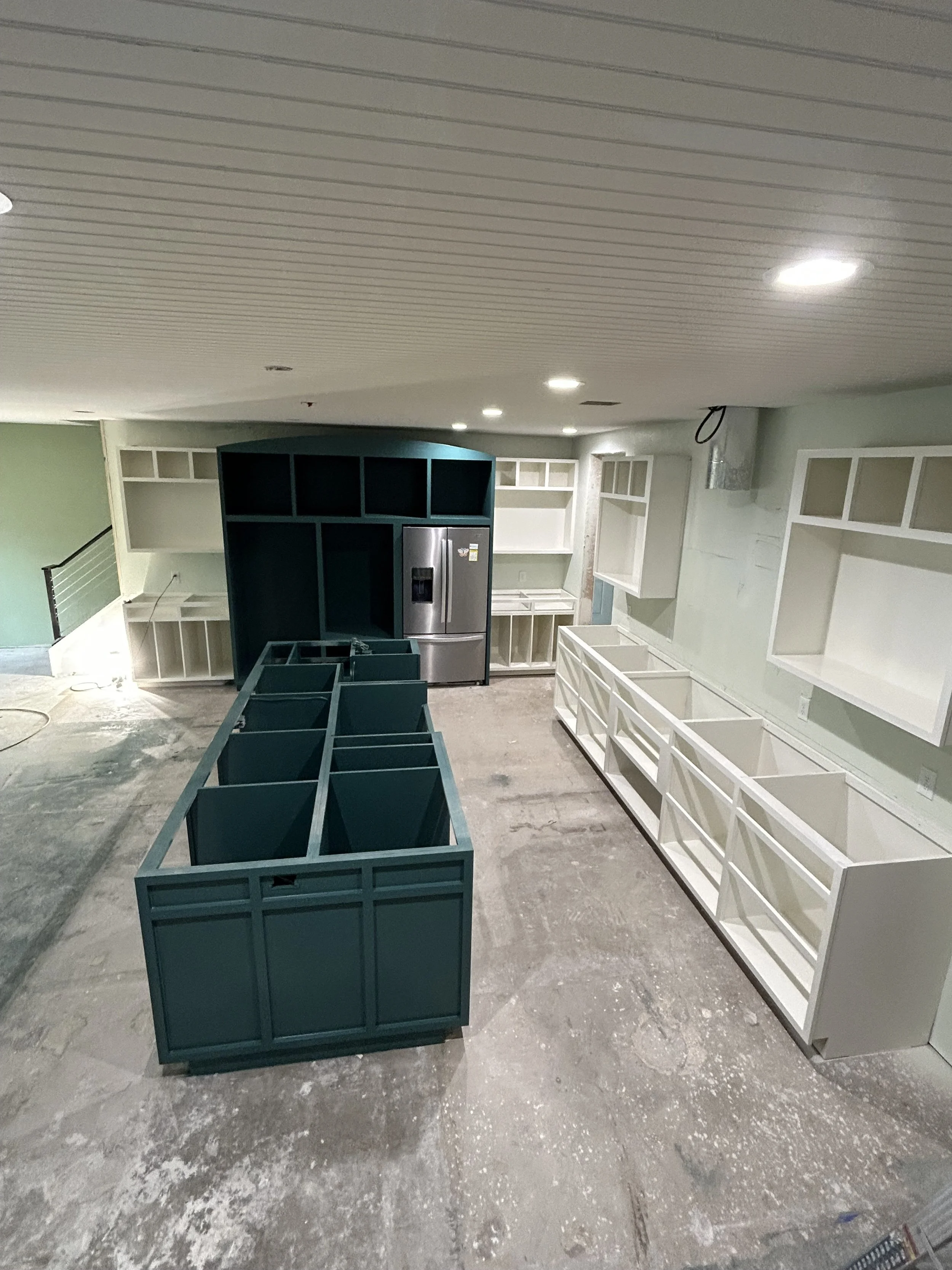 Unfinished kitchen with white open shelving, a stainless steel refrigerator, and a central blue cabinet with open compartments, on a concrete floor.