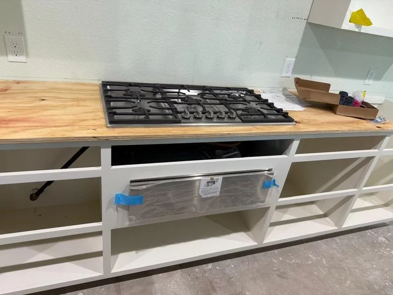 Kitchen countertop with a gas stove, a cardboard box, and some tools or supplies, with open shelving below and a light-colored wall behind.