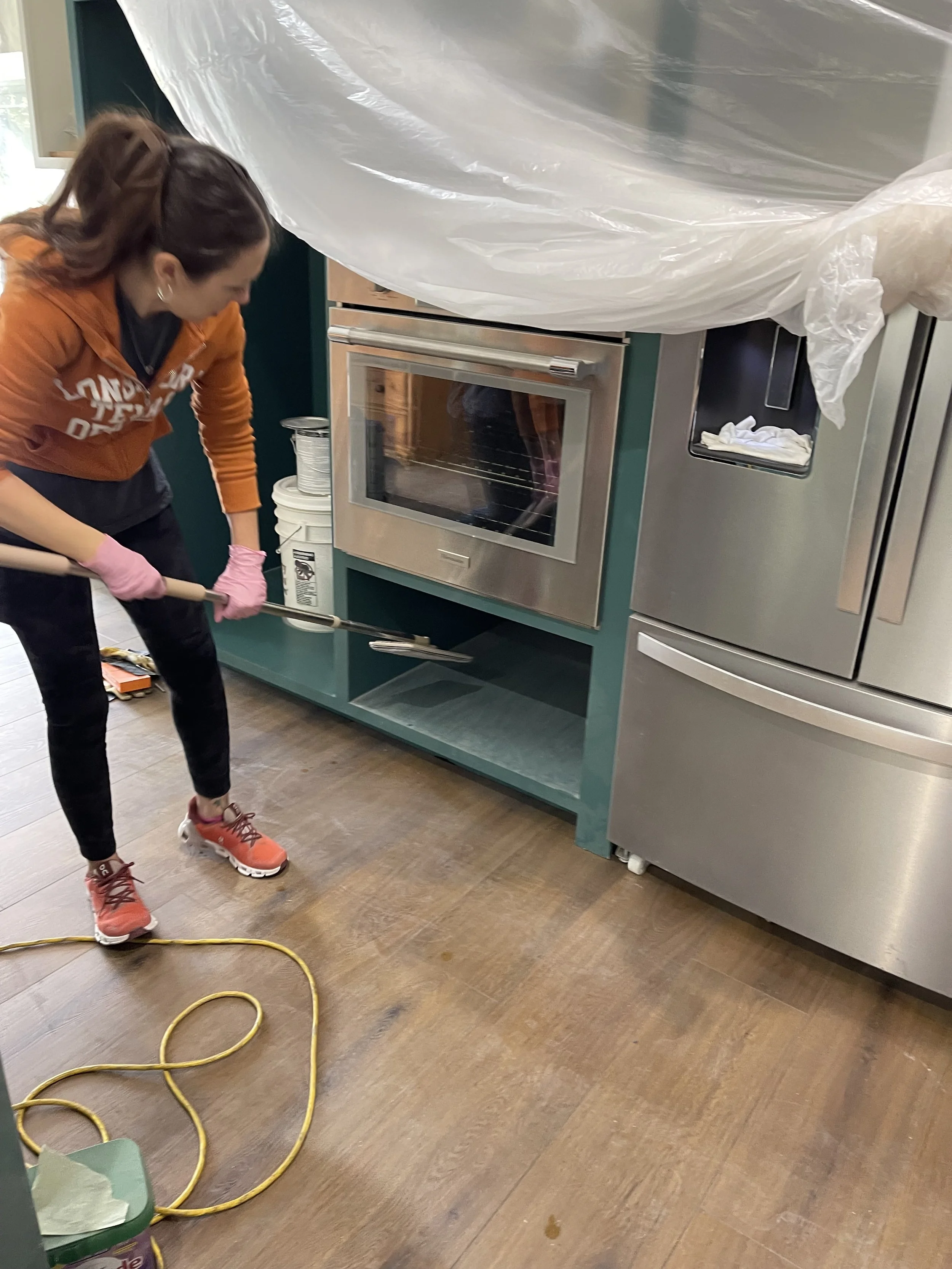 A woman cleaning a wooden floor in a kitchen with a yellow extension cord nearby. She is wearing a brown hoodie, black pants, pink gloves, and pink sneakers, holding a mop.