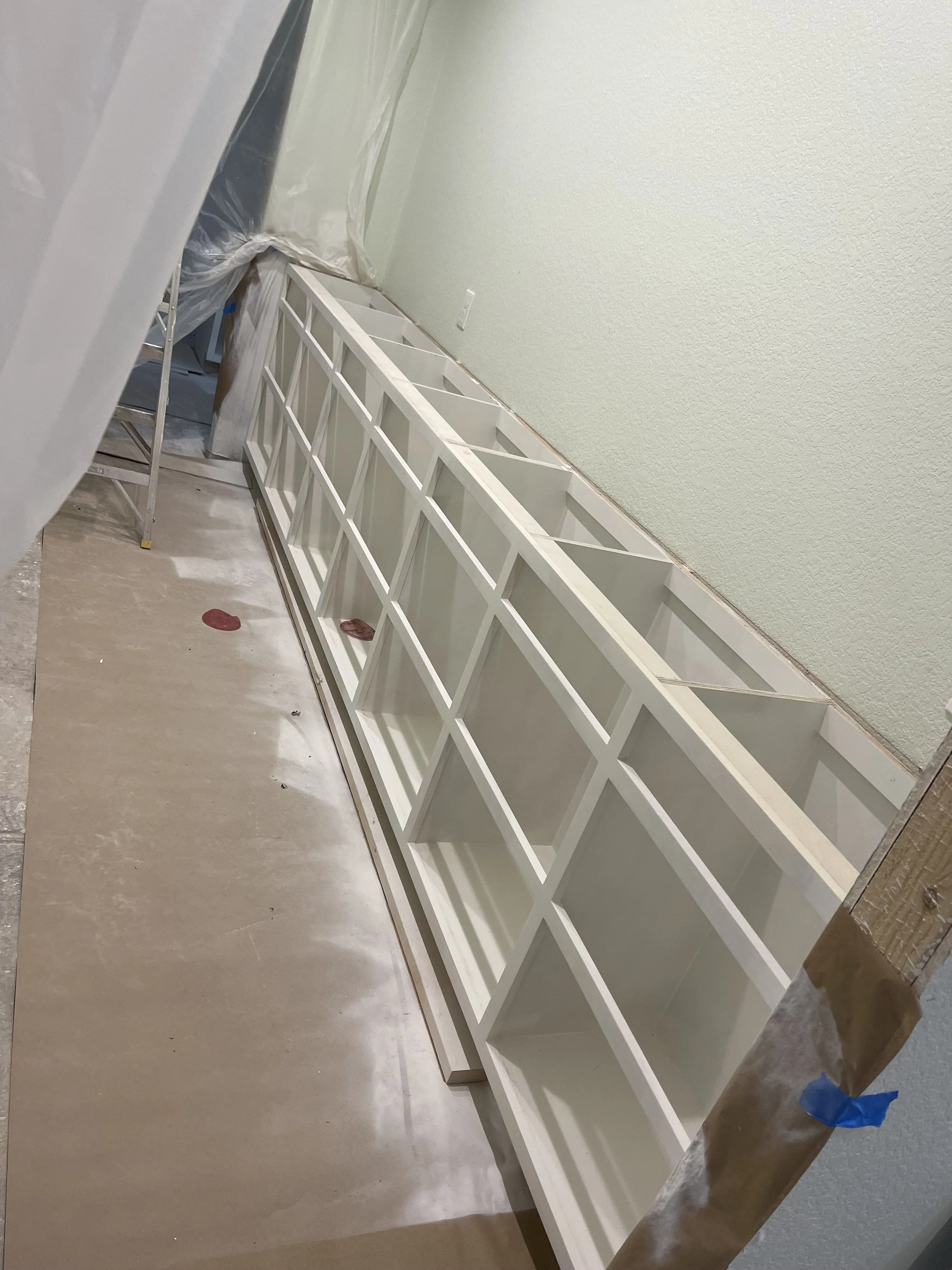 White built-in shelving unit being installed against a light green wall, with tools and protective coverings nearby in a room under construction.