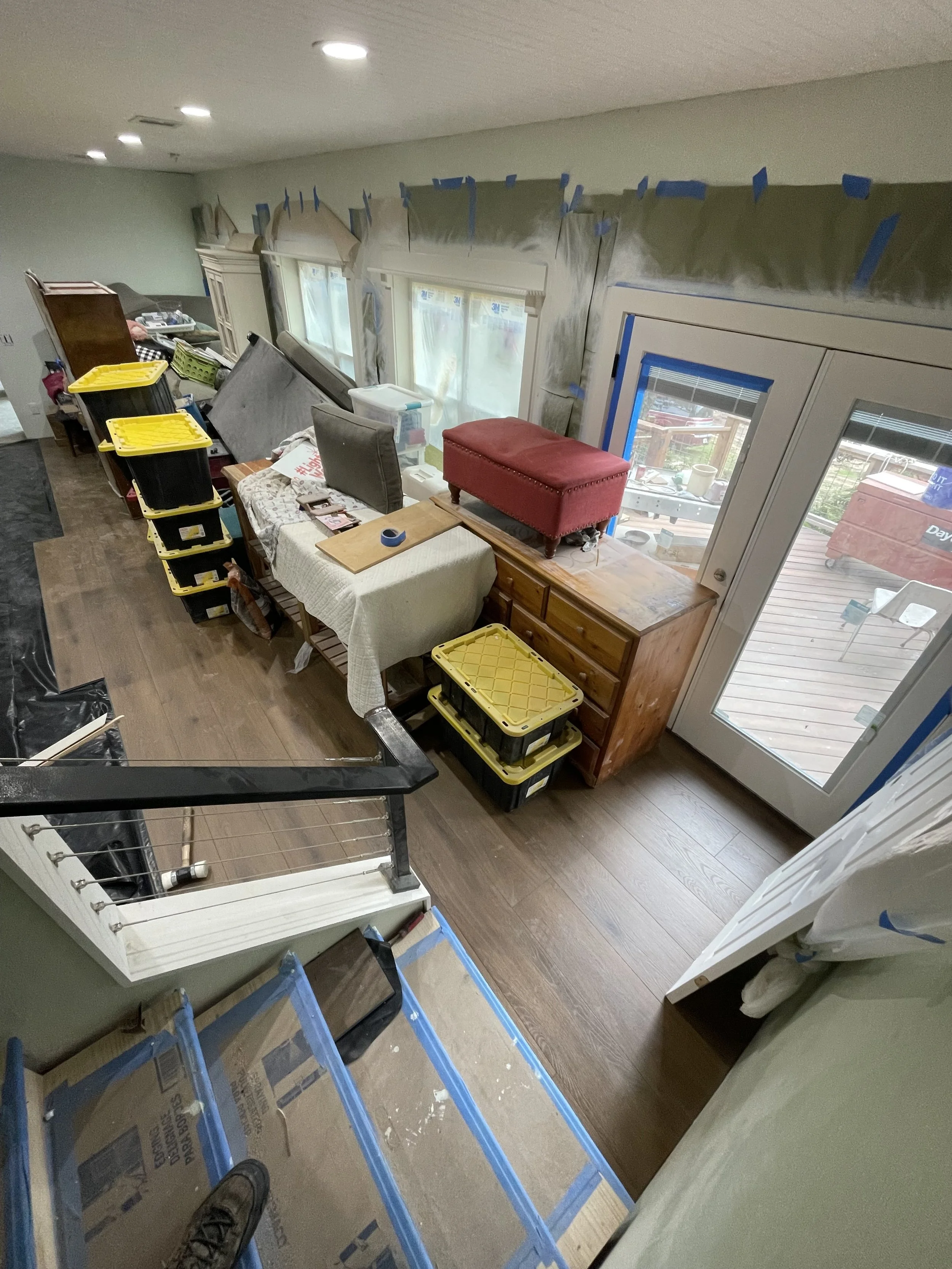 Room under renovation with plastic sheets, tools, chairs, and storage containers, also on a wooden floor near large windows and a glass door.