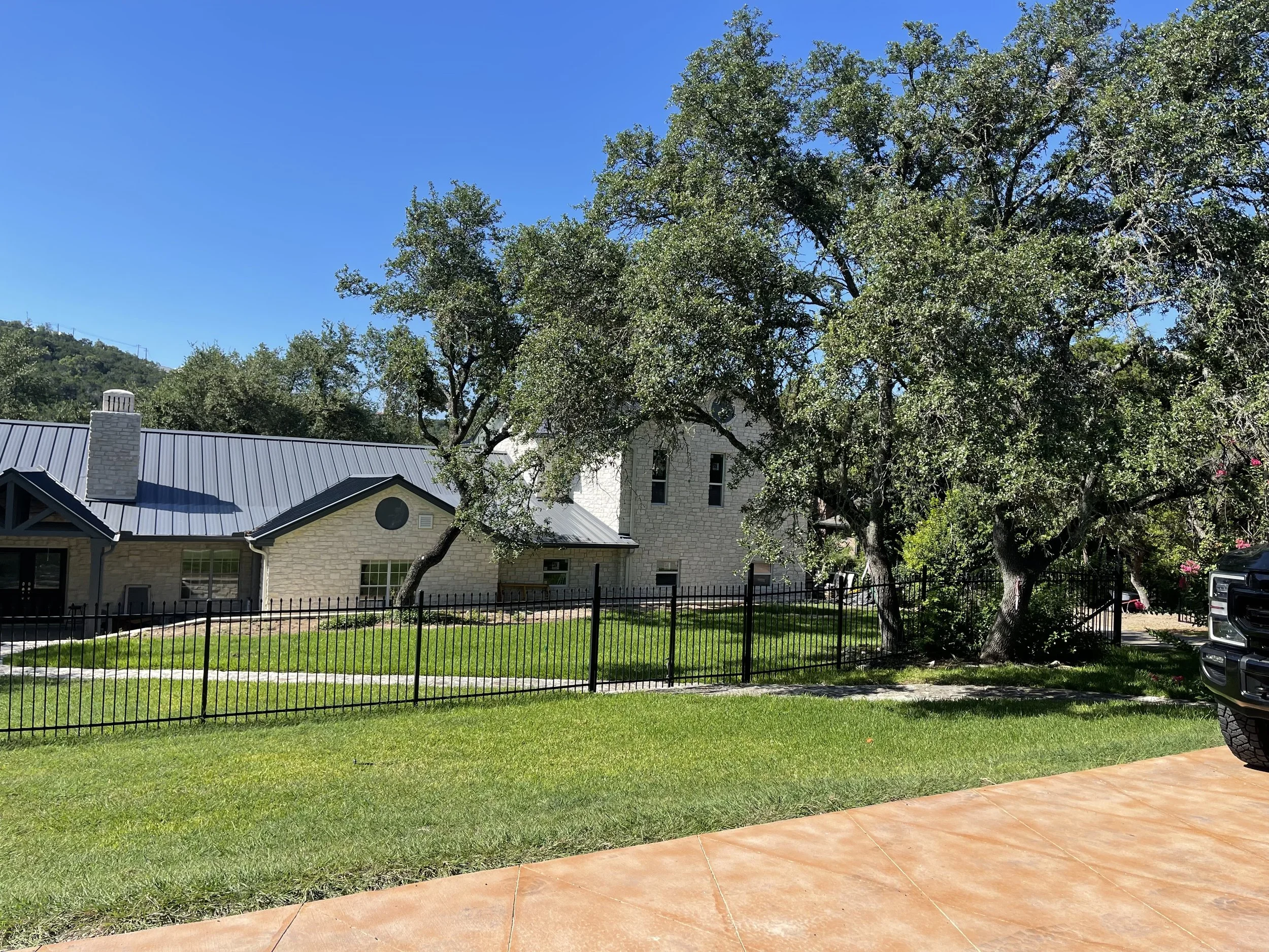View of a house with a metal roof, surrounded by trees and a grassy yard, with a parking lot and part of a black vehicle on the right side, under a clear blue sky.