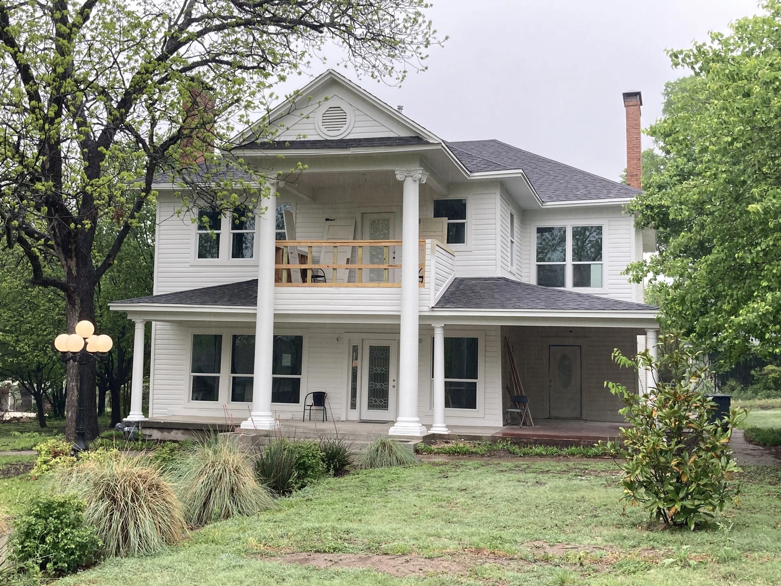 A two-story white house with a front porch and balcony, surrounded by green trees and grass.