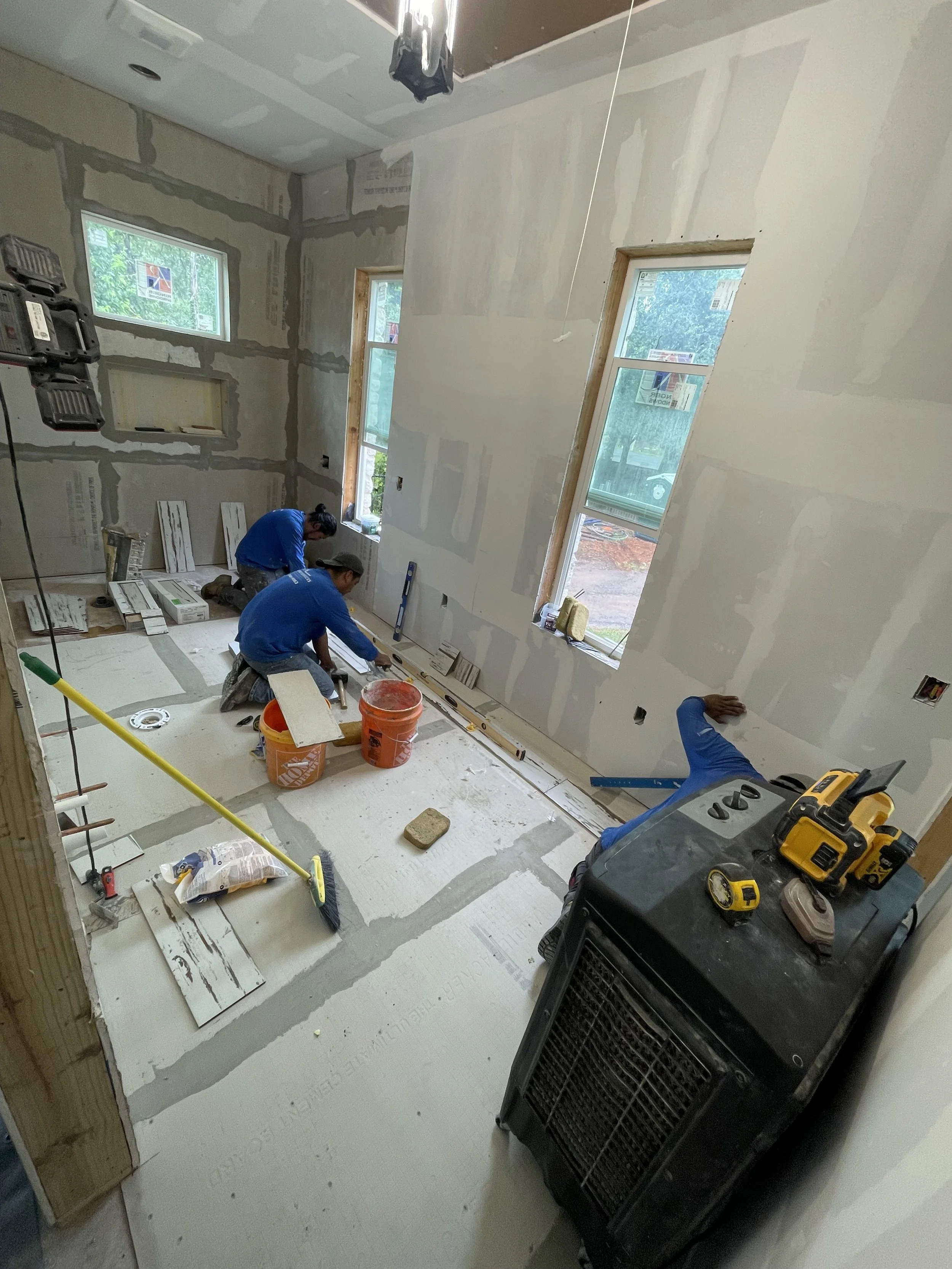 Three workers installing flooring in a room under construction, with drywall walls, two large windows, and construction tools and supplies around.