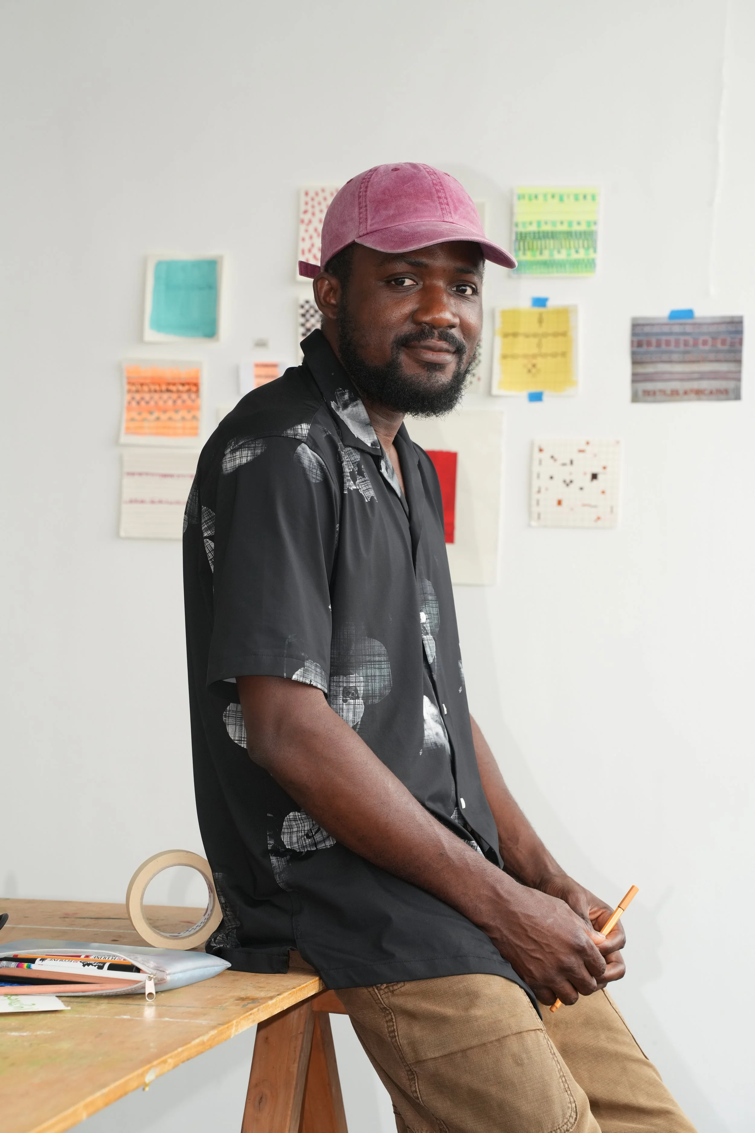 A man with a beard and a pink cap sitting on a wooden stool in front of an art wall, holding an orange pencil.