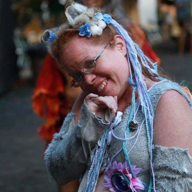 A woman with dreadlocks decorated with blue and gray yarn, wearing glasses and a cozy gray top, smiling and making a playful gesture at a lively outdoor event.