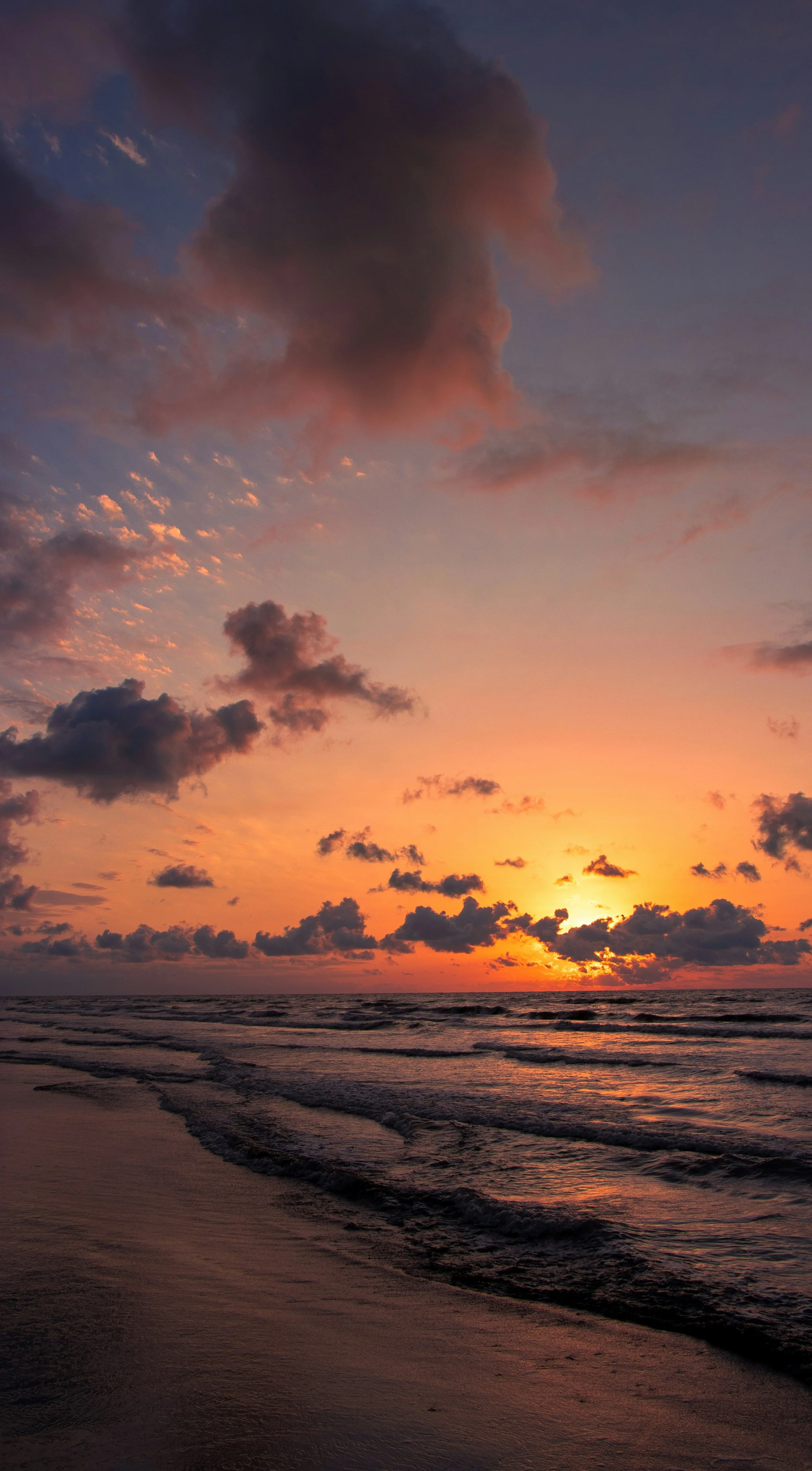 Sunset over the ocean with a colorful sky and clouds, waves gently hitting the sandy shore.