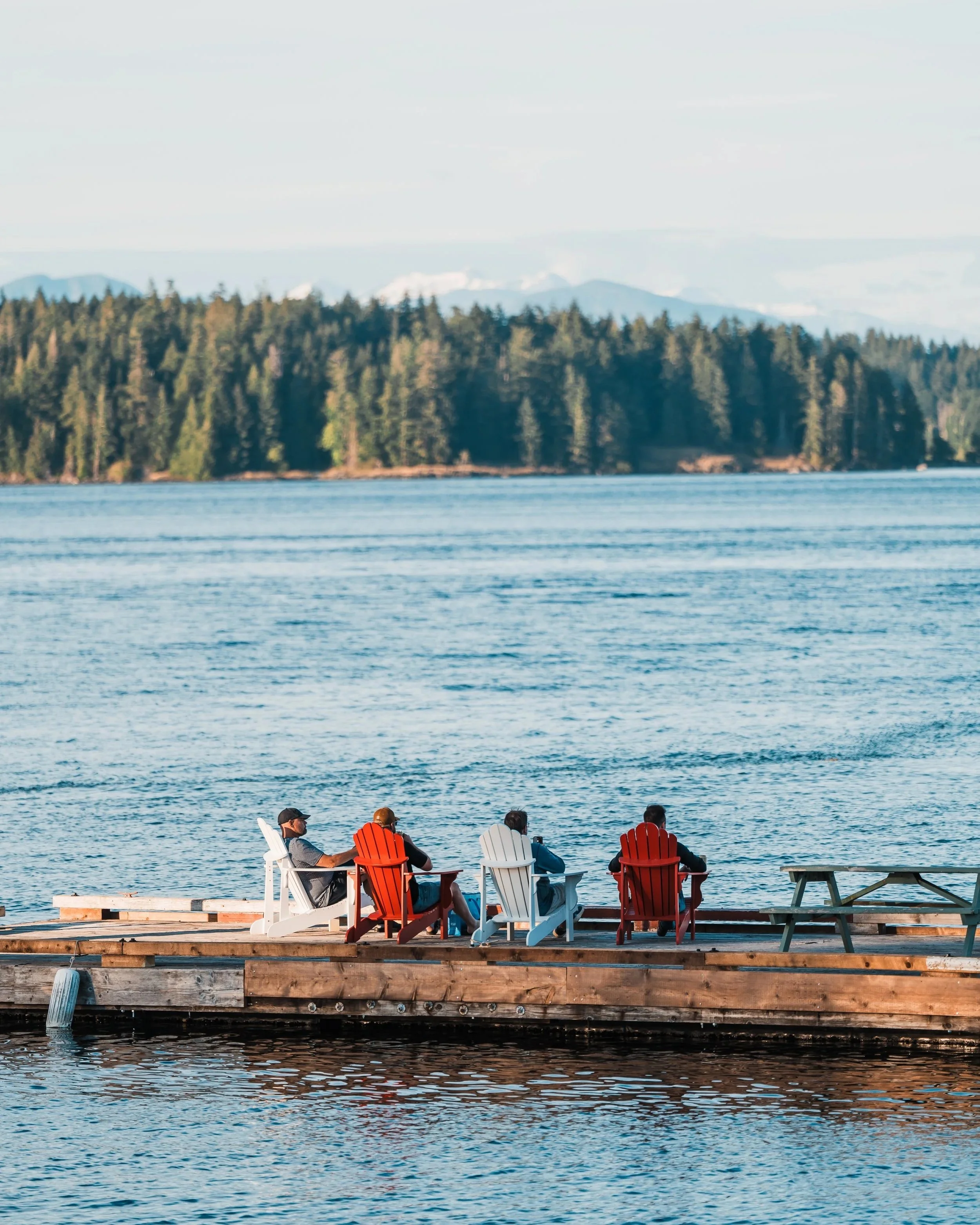 Four people sitting on colorful Adirondack chairs on a wooden dock by a body of water, with a forested shoreline and mountains in the background.