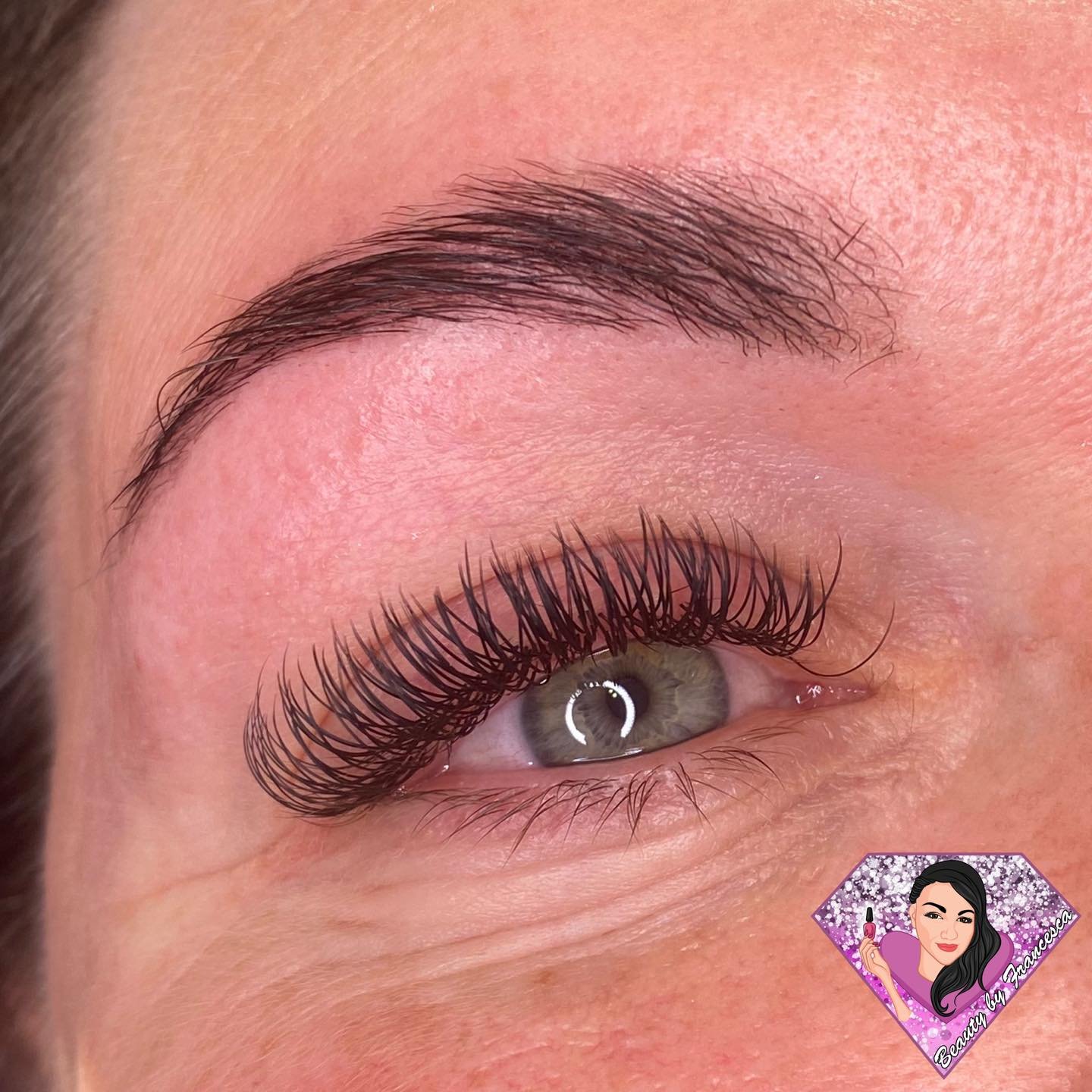 Close-up of a woman's eye with green iris, natural makeup, and eyelash extensions, showing the eyebrow and surrounding skin.