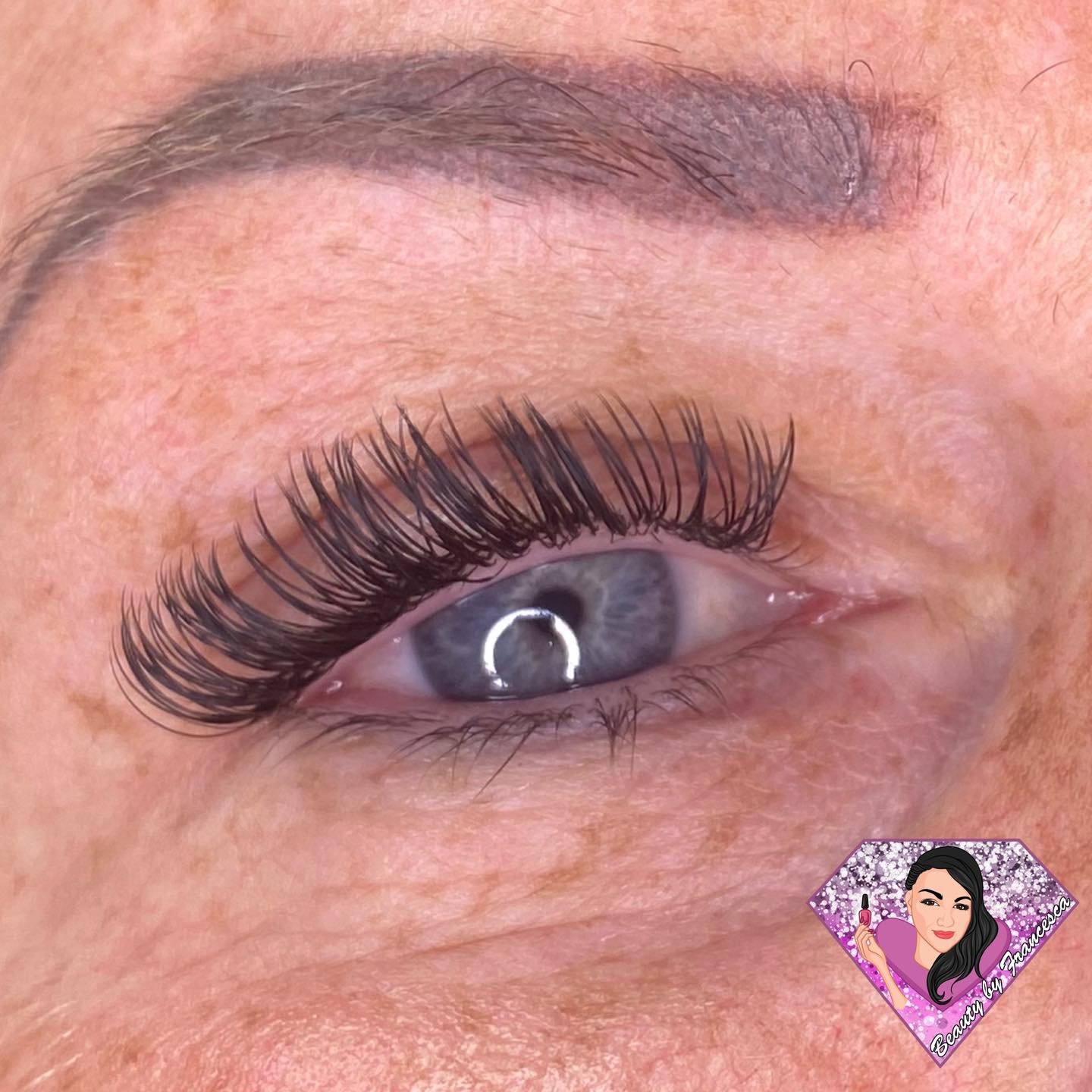 A close-up of a woman's eye with long, thick, curled eyelash extensions, highlighting her gray-blue iris and eyebrow.