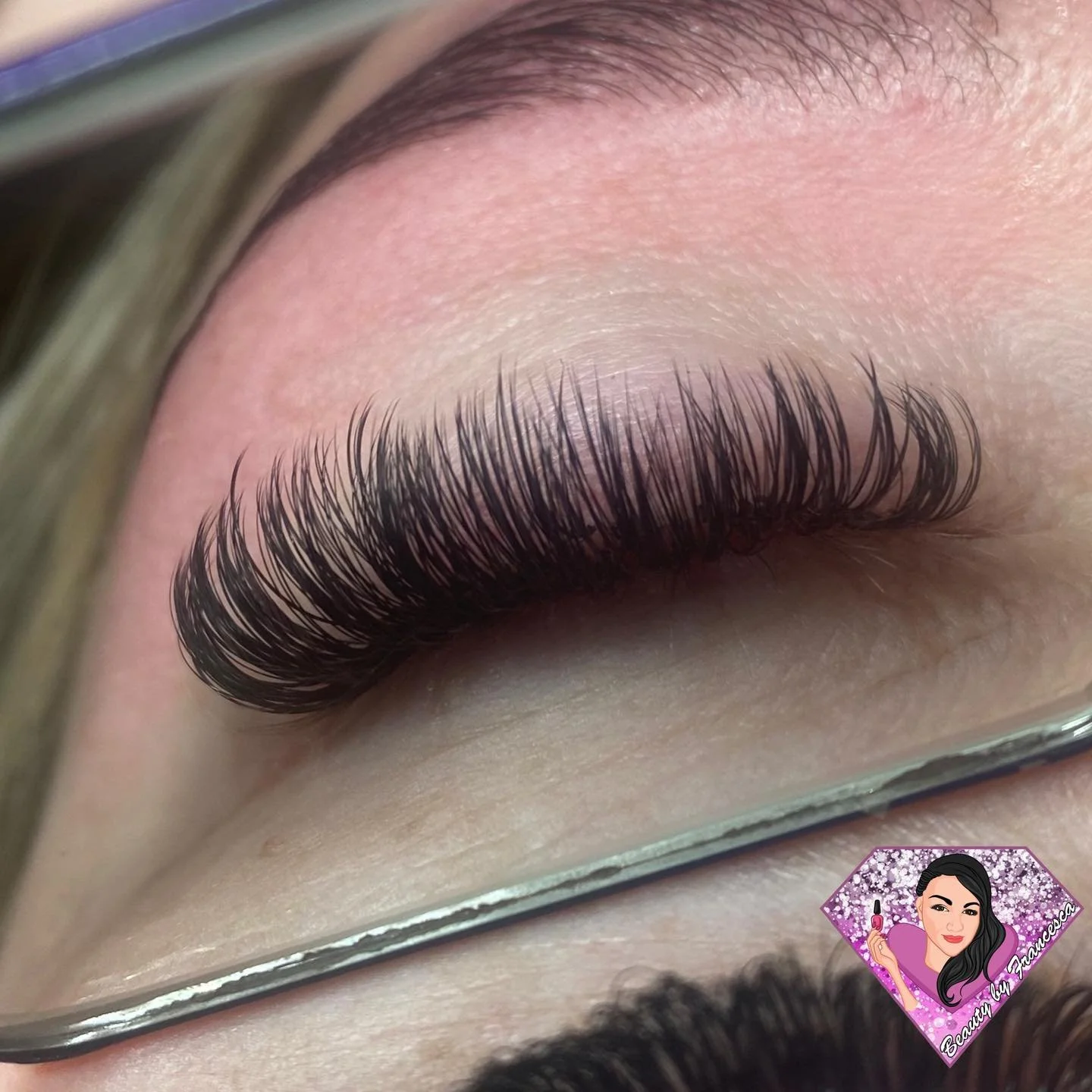Close-up of a closed eye with long, thick, curled eyelashes, shaved eyebrows, and light skin. A mirror is visible below the eye reflection.