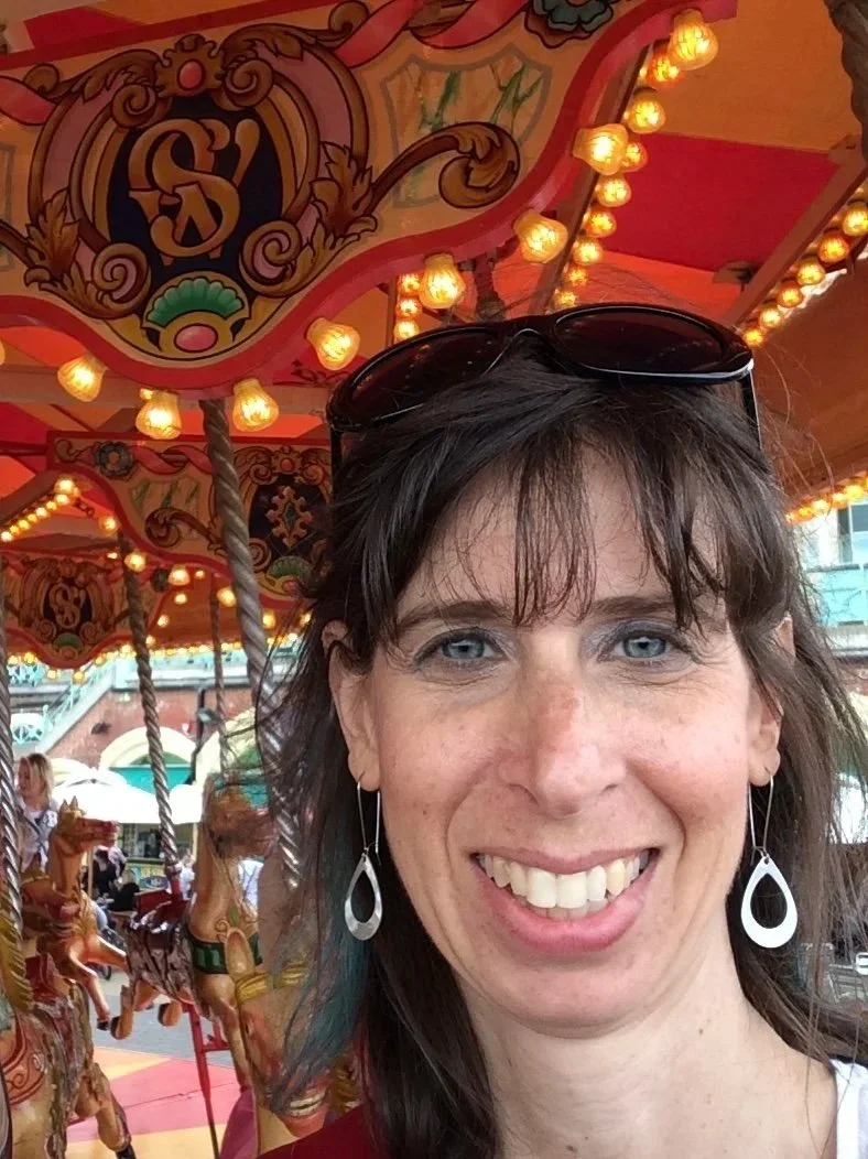 a photo of Amy - a white woman with shoulder length brown hair, wearing black sunglasses on top of her head and long dangling silver earrings. She is outdoors on a carousel. 