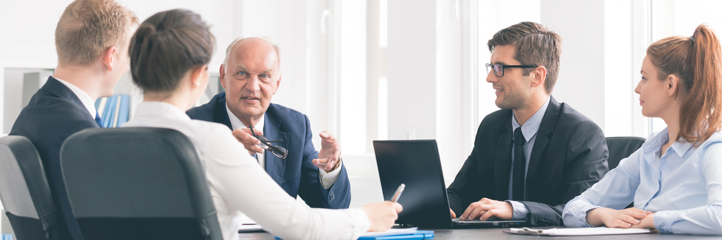 A business meeting with five professionals sitting around a table in a bright office, engaged in a discussion with an older man speaking and the others listening.