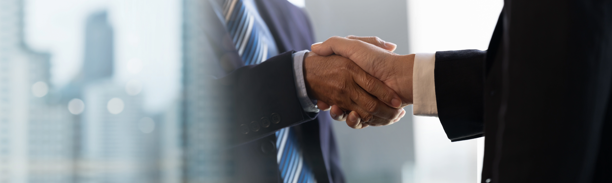 Close-up of two people shaking hands in a professional setting, with one person wearing a dark suit and a striped tie, and the other person wearing a black suit and white shirt.  Strategic Alignment.