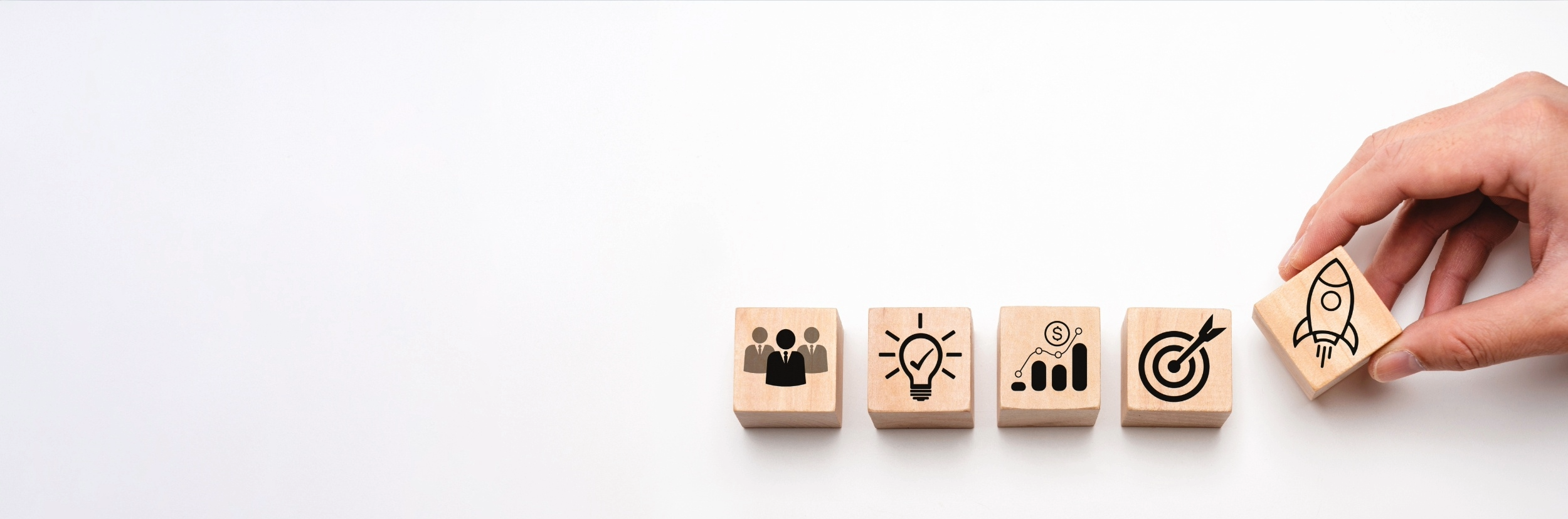 A hand placing a wooden block with a rocket icon next to other blocks with icons of a group of people, a lightbulb, a bar graph with dollar sign, and a target with an arrow, on a white surface.