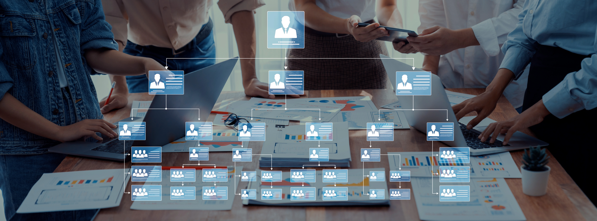 A group of people gathered around a conference table reviewing documents and working on laptops with a digital overlay of a hierarchical organizational chart.