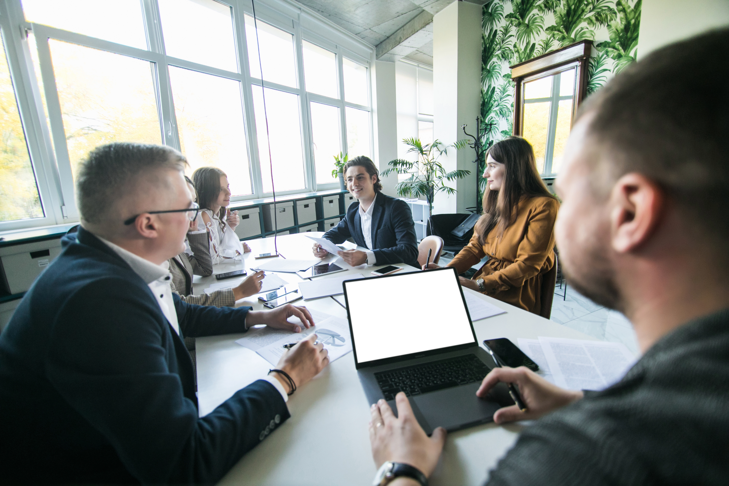 Six people sitting around a white conference table in a bright room with large windows, having a business meeting.