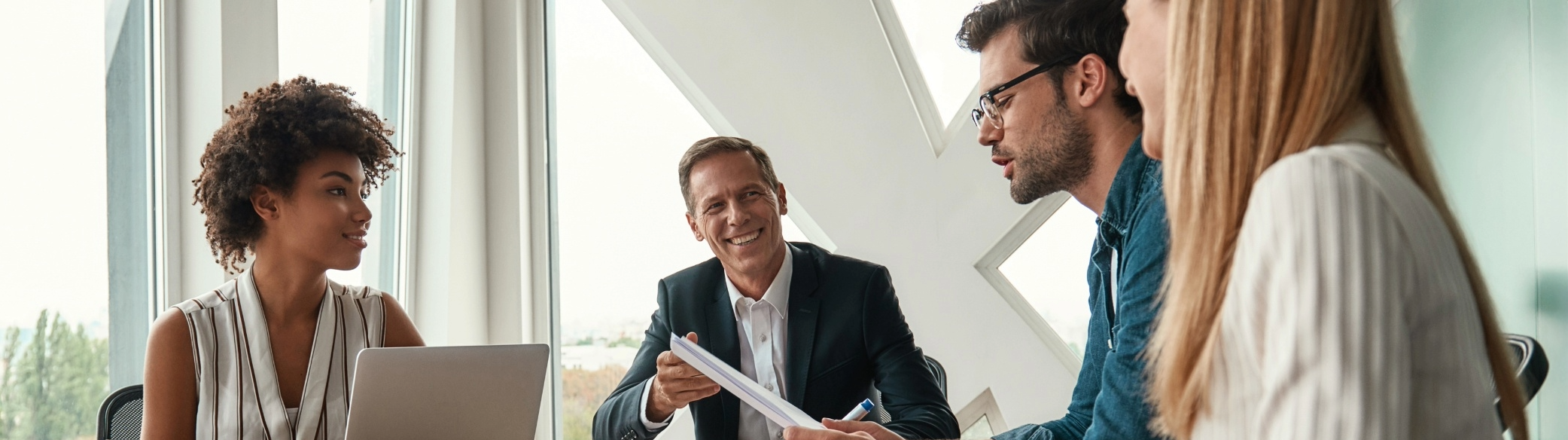 A diverse group of four professionals engaged in a discussion in a modern conference room with large windows, with a man in a suit smiling and holding a notebook.