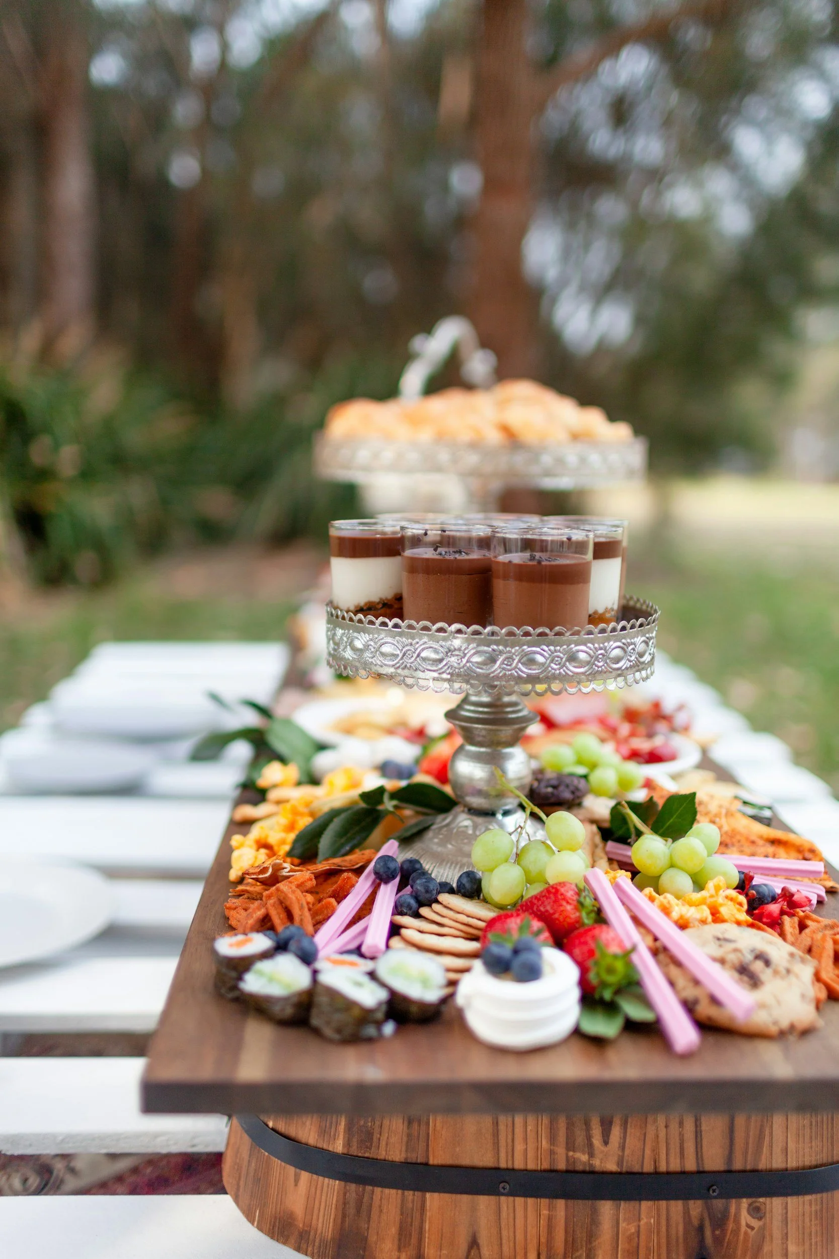 A dessert table outdoors with assorted sweets and fruit, topped with chocolate mousse and surrounded by grapes and cookies.