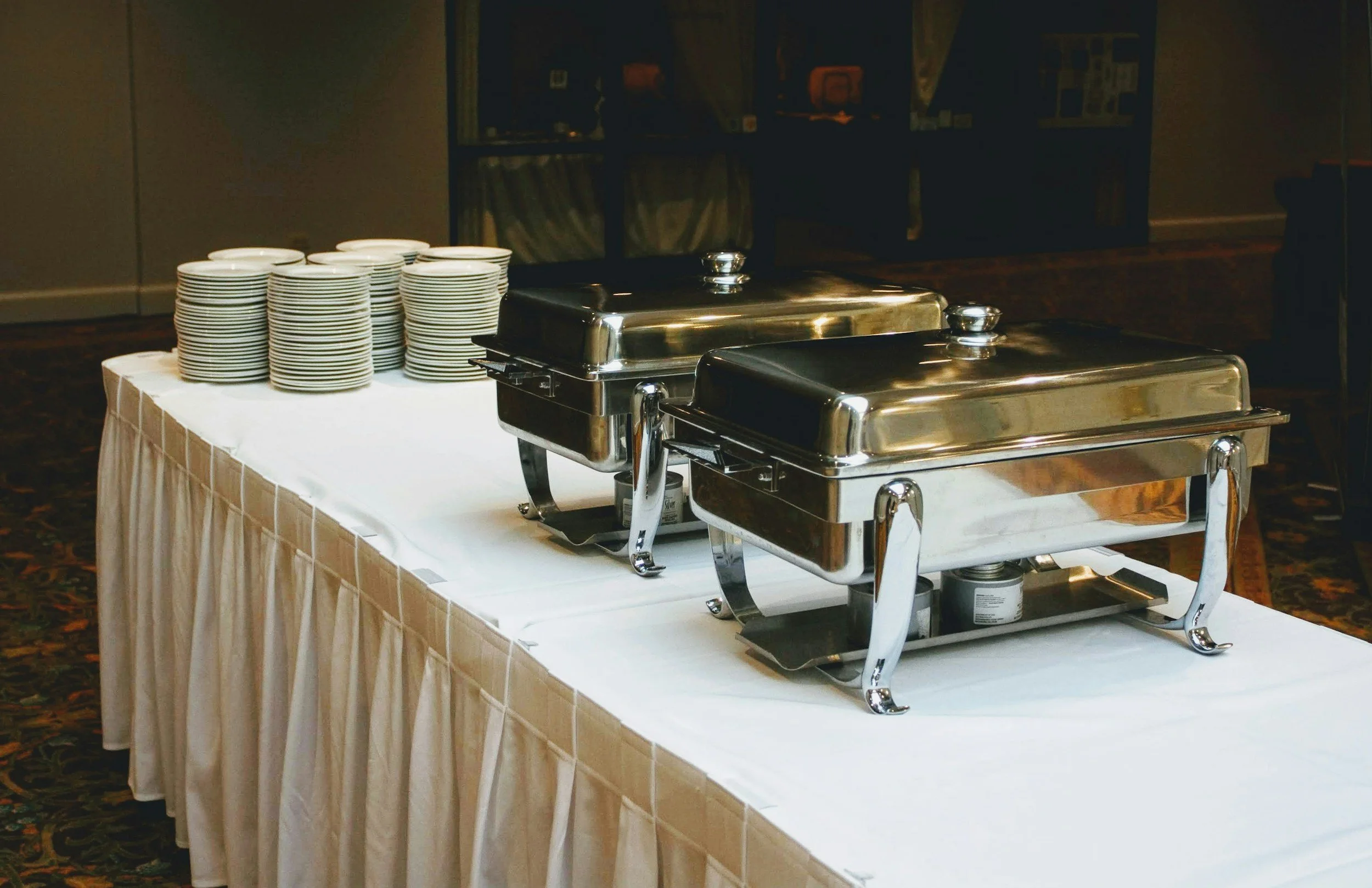 Chafing dishes and stacks of plates on a table set up for a buffet.