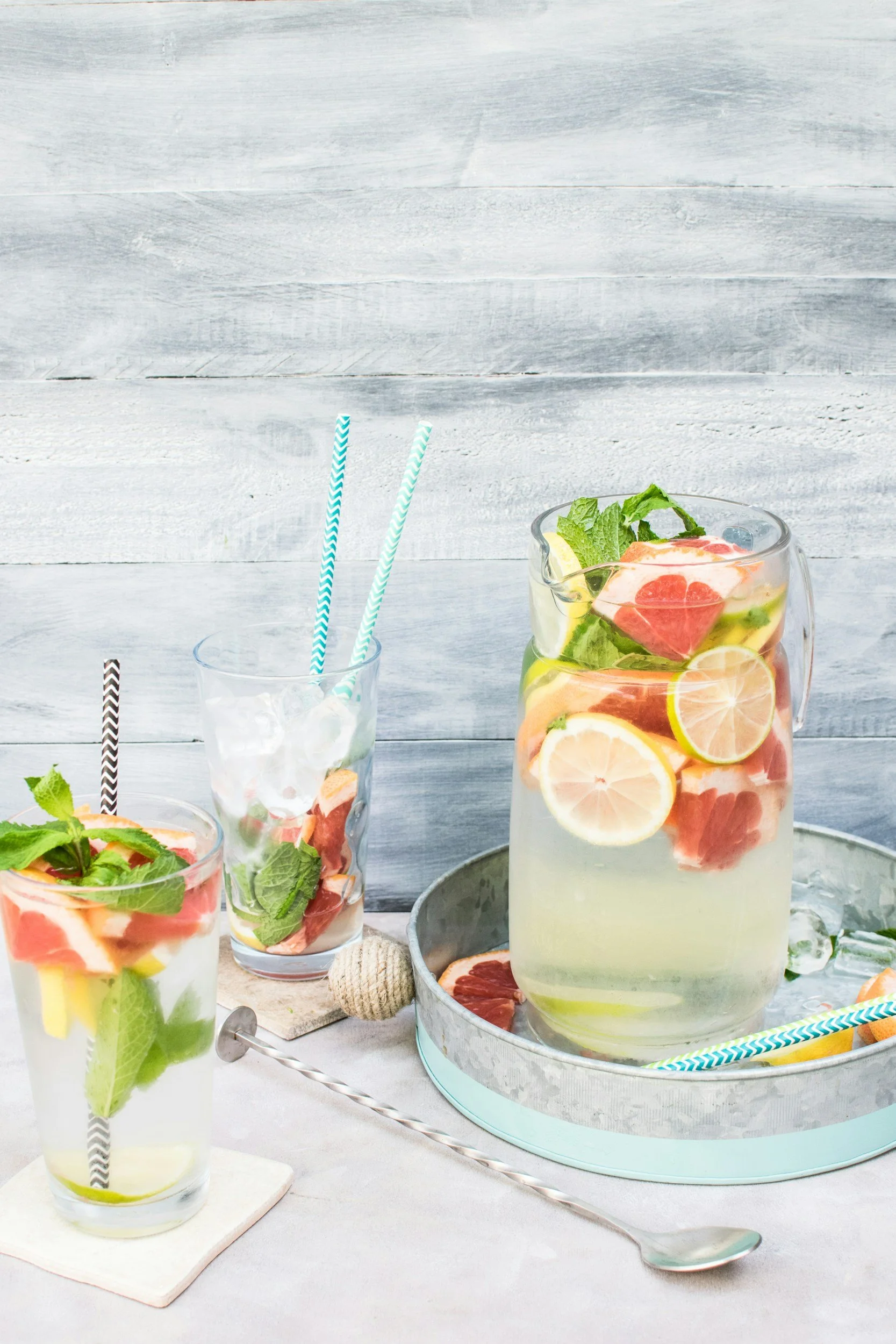A glass pitcher and two tall glasses filled with fruit-infused water, garnished with mint leaves, lemon, lime, grapefruit, and watermelon slices, set on a light-colored surface with a gray wooden background.