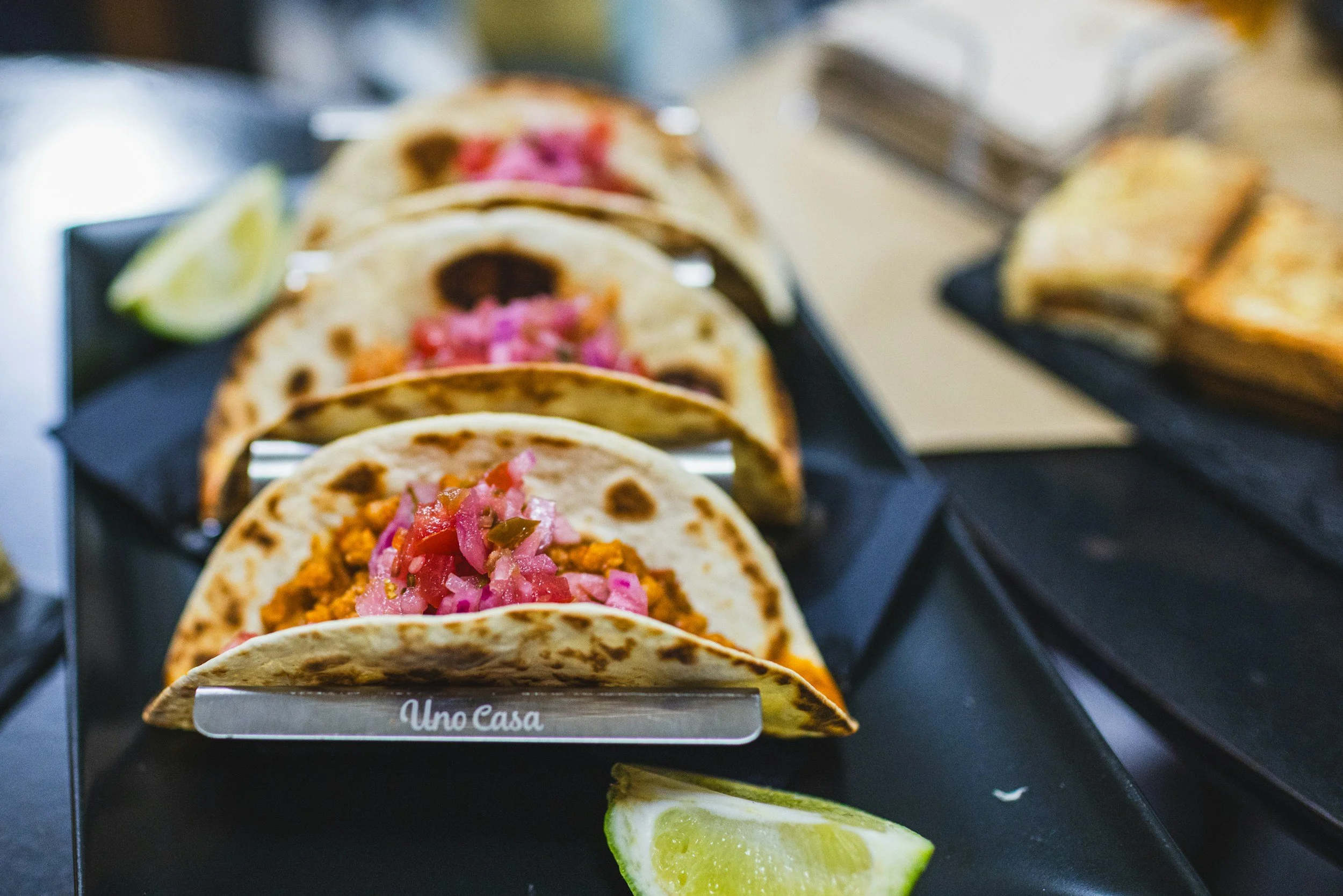 Close-up of three tacos filled with seasoned meat, topped with chopped pickled red onions, served on a black plate with lime wedges on the side.