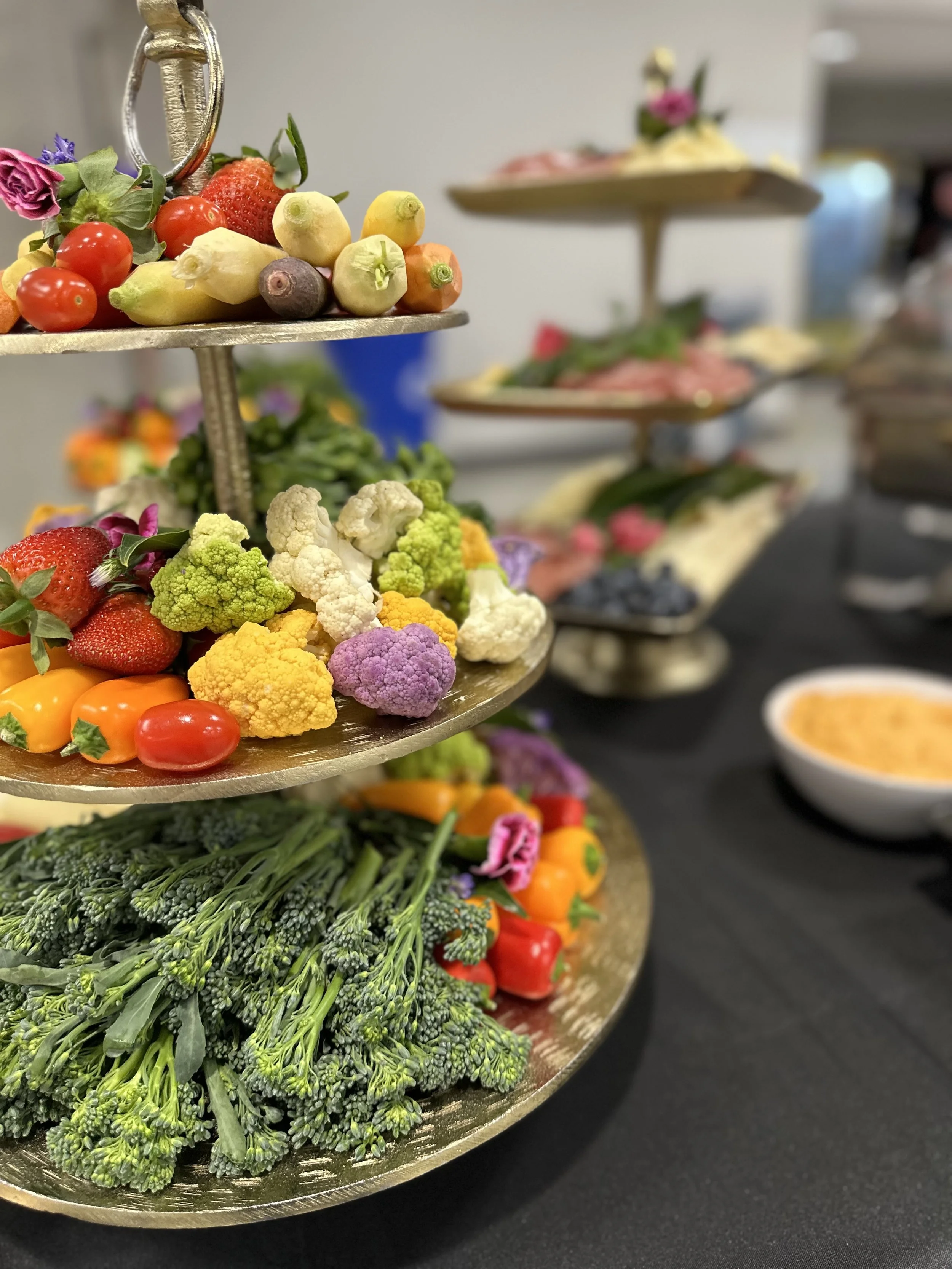 Tiered display of colorful fresh vegetables and fruits, including cauliflower, broccoli, cherry tomatoes, strawberries, peppers, and fingerling potatoes, on a table with a black tablecloth.