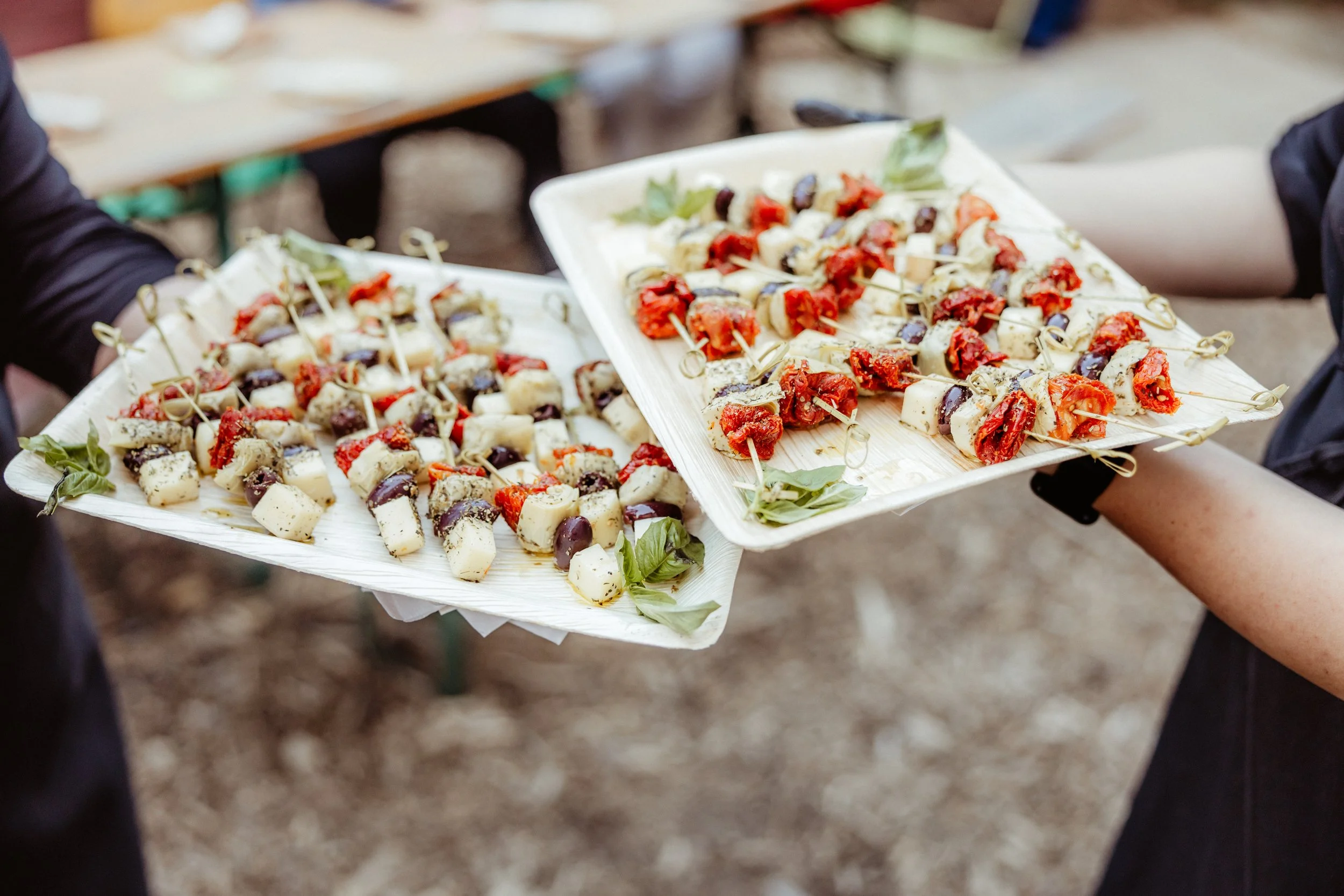 Two people holding trays with skewered appetizers, including cheese, sun-dried tomatoes, olives, and basil leaves.