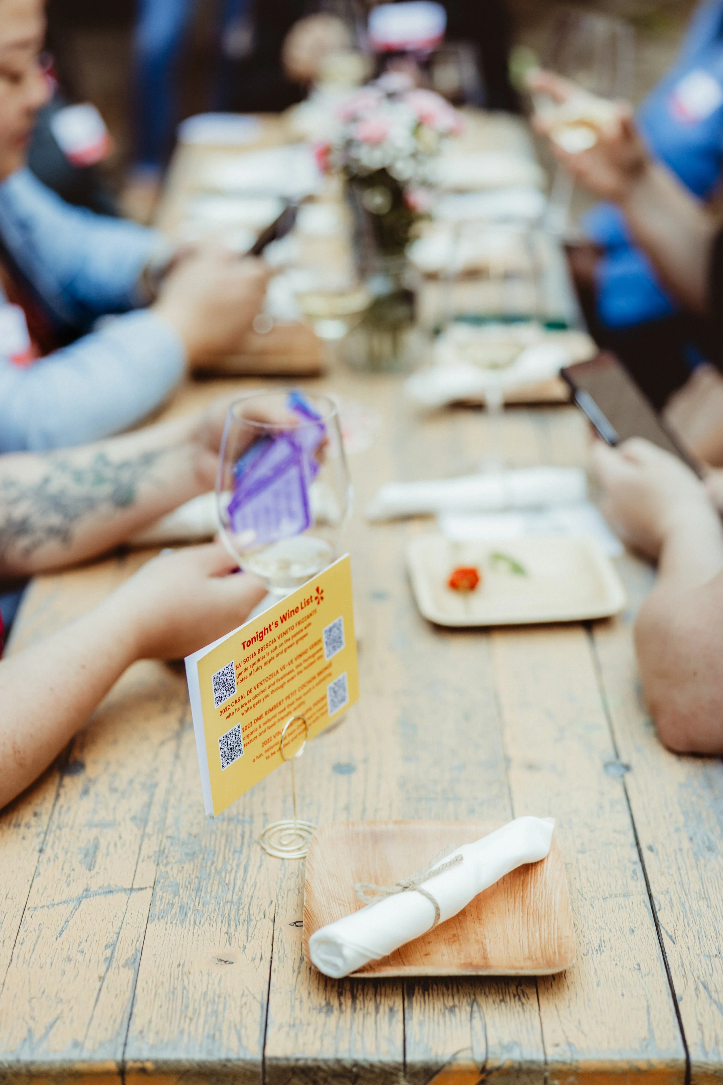 A group of people sitting at a wooden table with wine glasses, plates, and a menu card, during a social gathering.