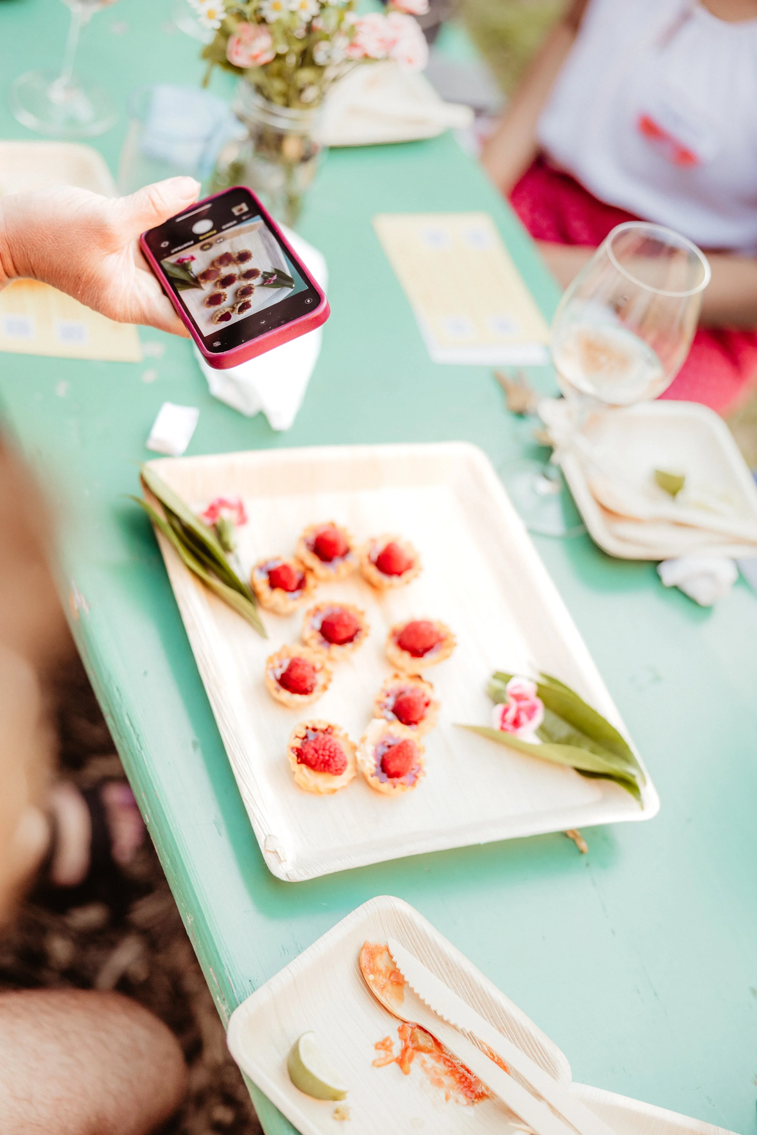 Person taking a photo of small desserts with raspberry toppings on a white tray at a table with glasses, plates, and floral decorations.