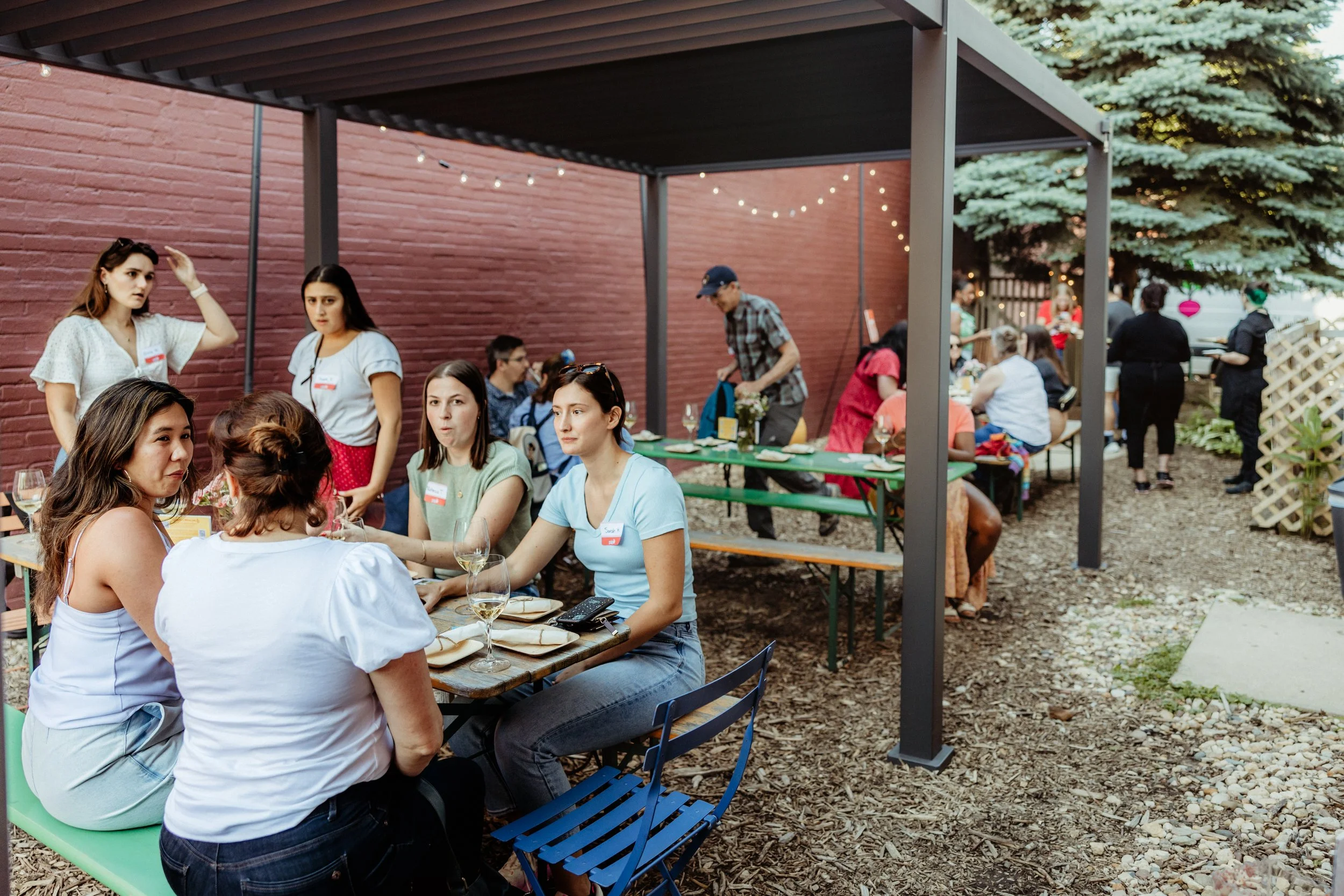 Group of people dining outdoors under a shaded patio with string lights, surrounded by a brick wall and trees.