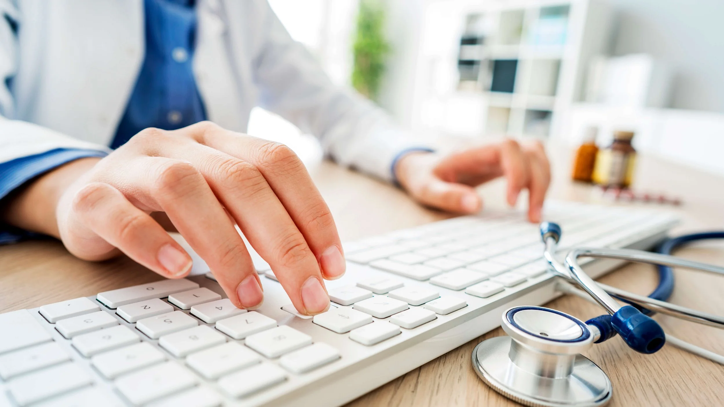 A healthcare professional's hand using a computer keyboard with a stethoscope on a wooden desk in a medical office.