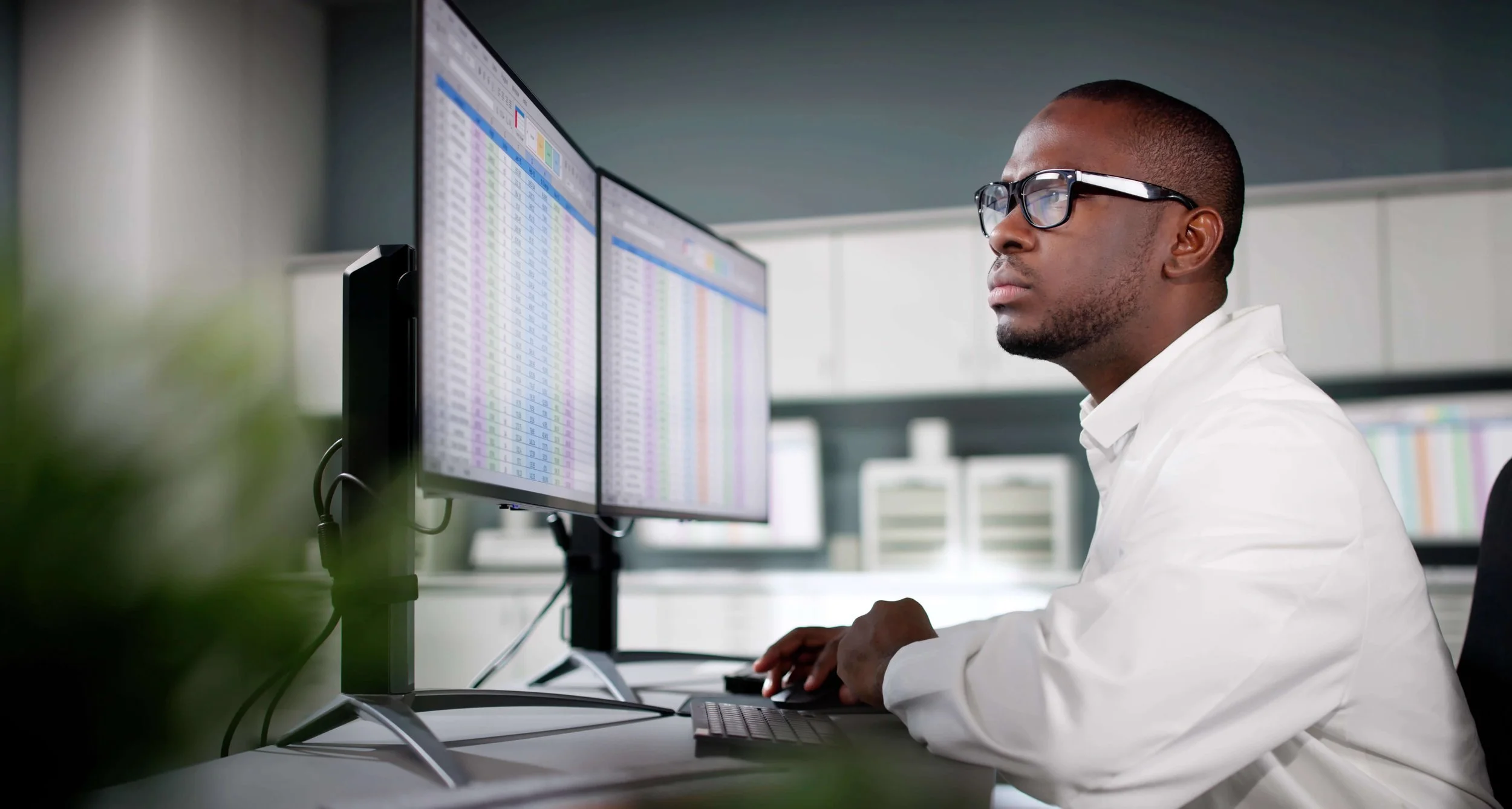 A man working at a desk with two computer monitors displaying colorful spreadsheets, wearing glasses and a white shirt.