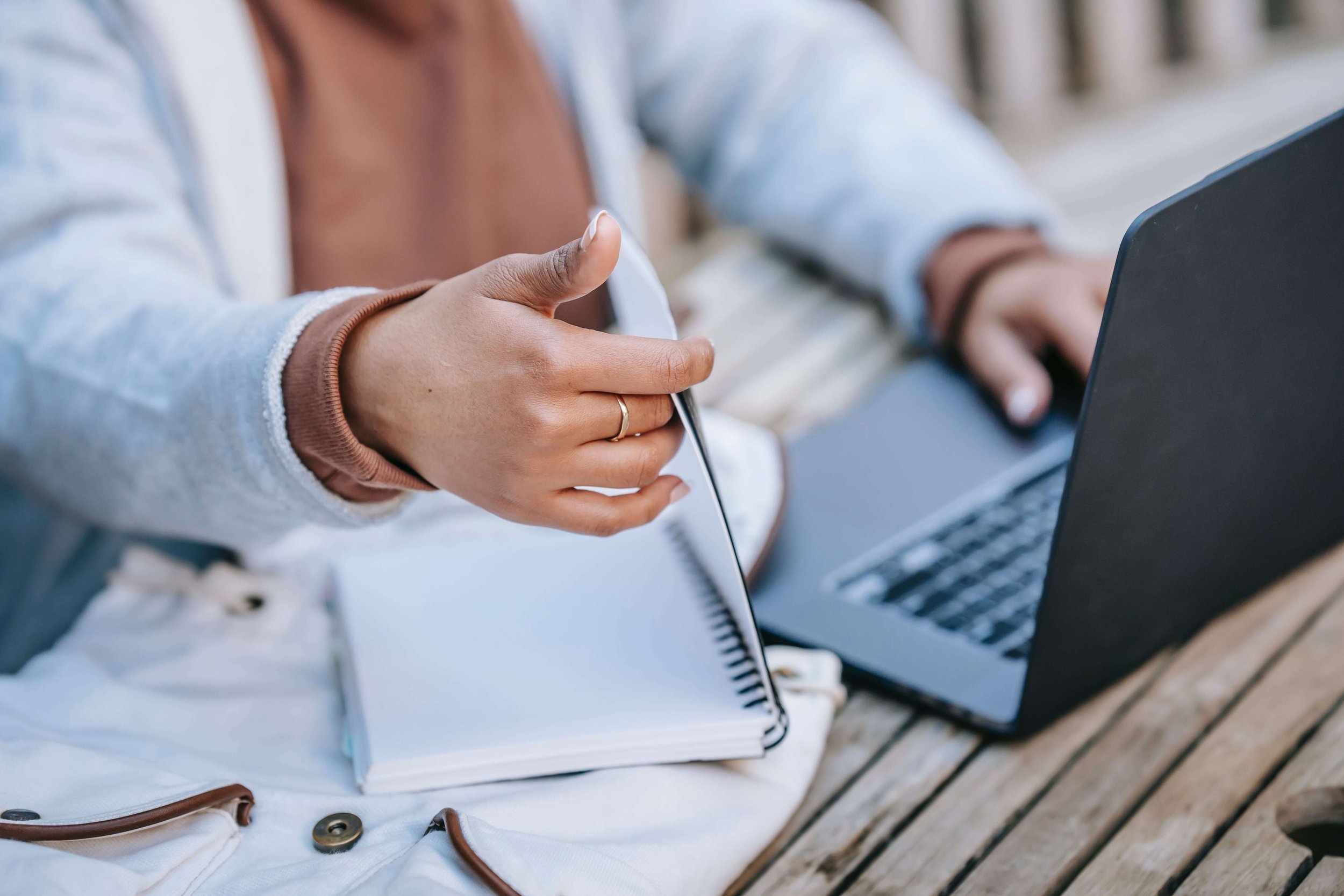 A person using a laptop and pointing at a notebook on a wooden table.
