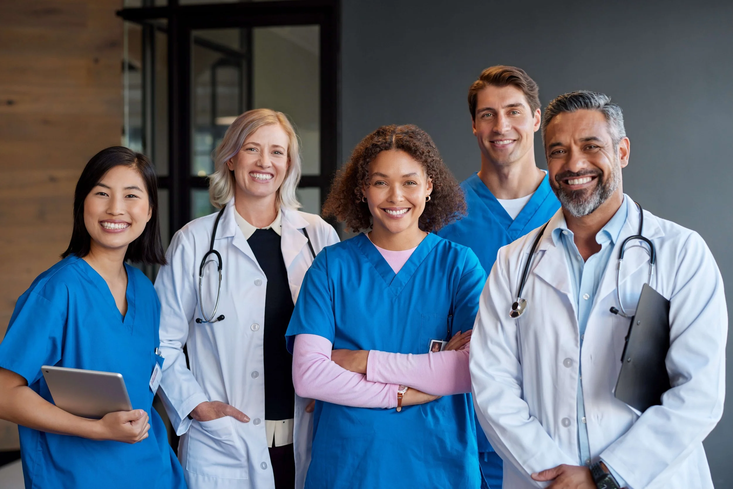 Group of five diverse healthcare professionals smiling, wearing medical scrubs and white coats, standing together in a hospital or clinic.