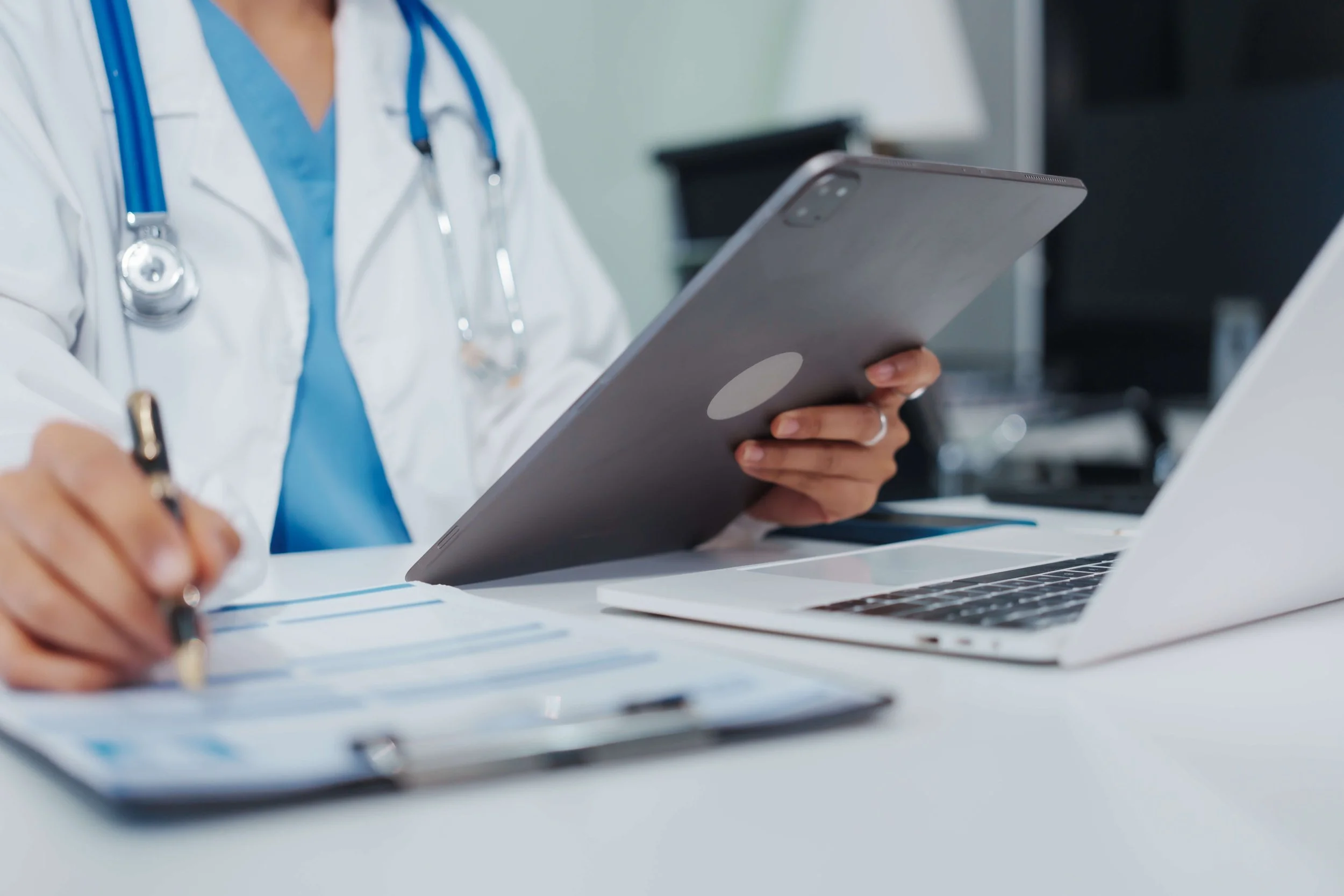 Medical professional wearing a white coat and stethoscope working at a desk with a laptop, tablet, notepad, and pen.