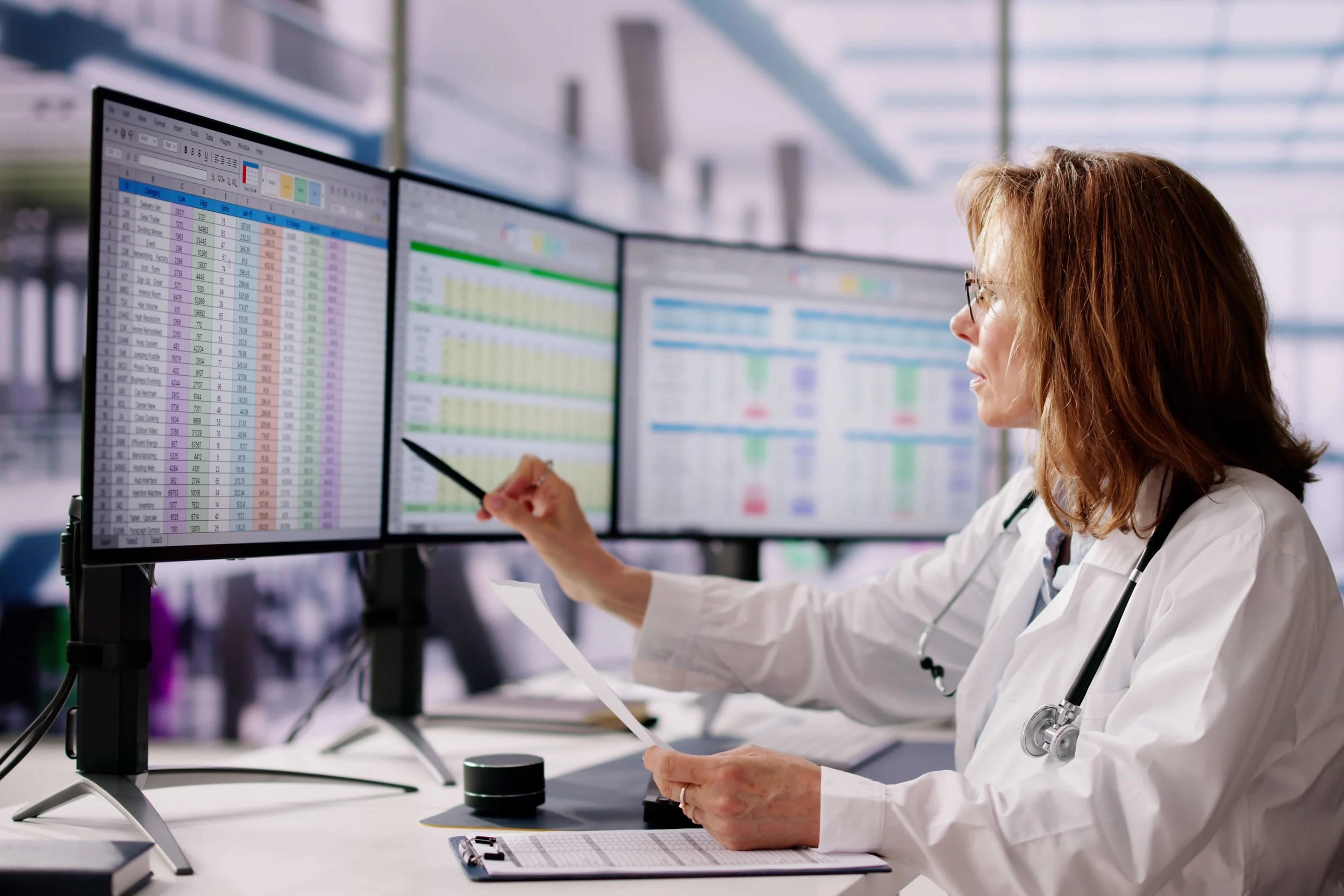 A female doctor or medical professional in a white coat, sitting at a desk with multiple computer monitors displaying spreadsheets and data, pointing at the screens with a pen.
