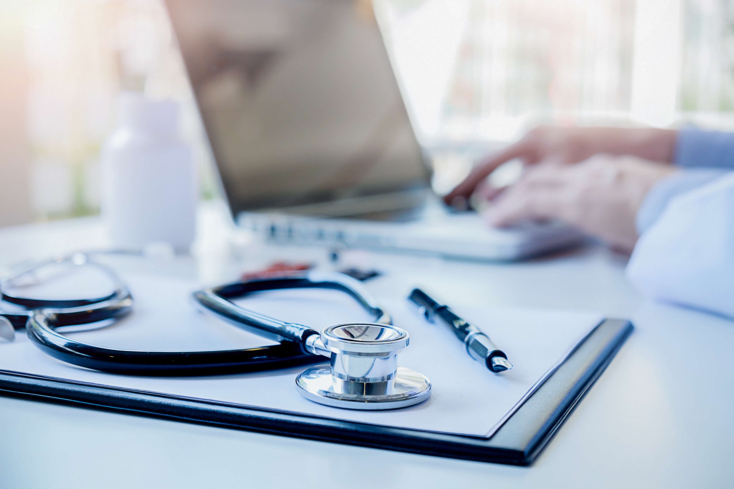 Medical stethoscope, pen, and clipboard on a white surface in a clinical setting, with a person typing on a laptop in the background.