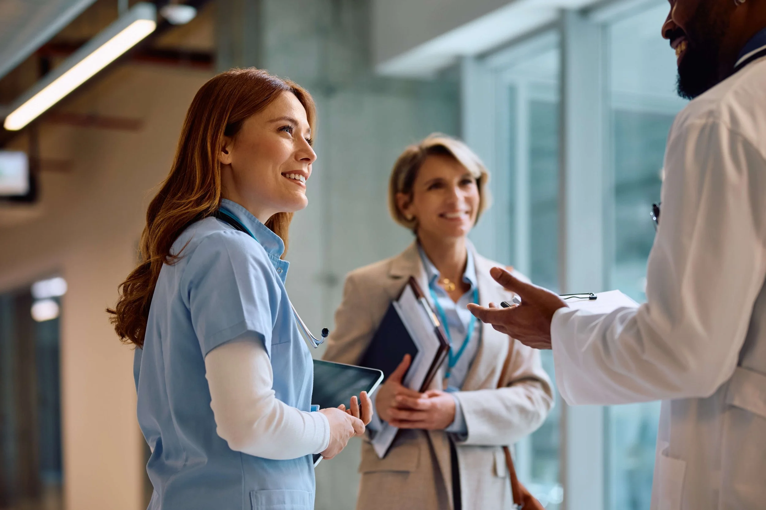 Three healthcare professionals in conversation in a hospital corridor, two women with notebooks and a man in a white coat.