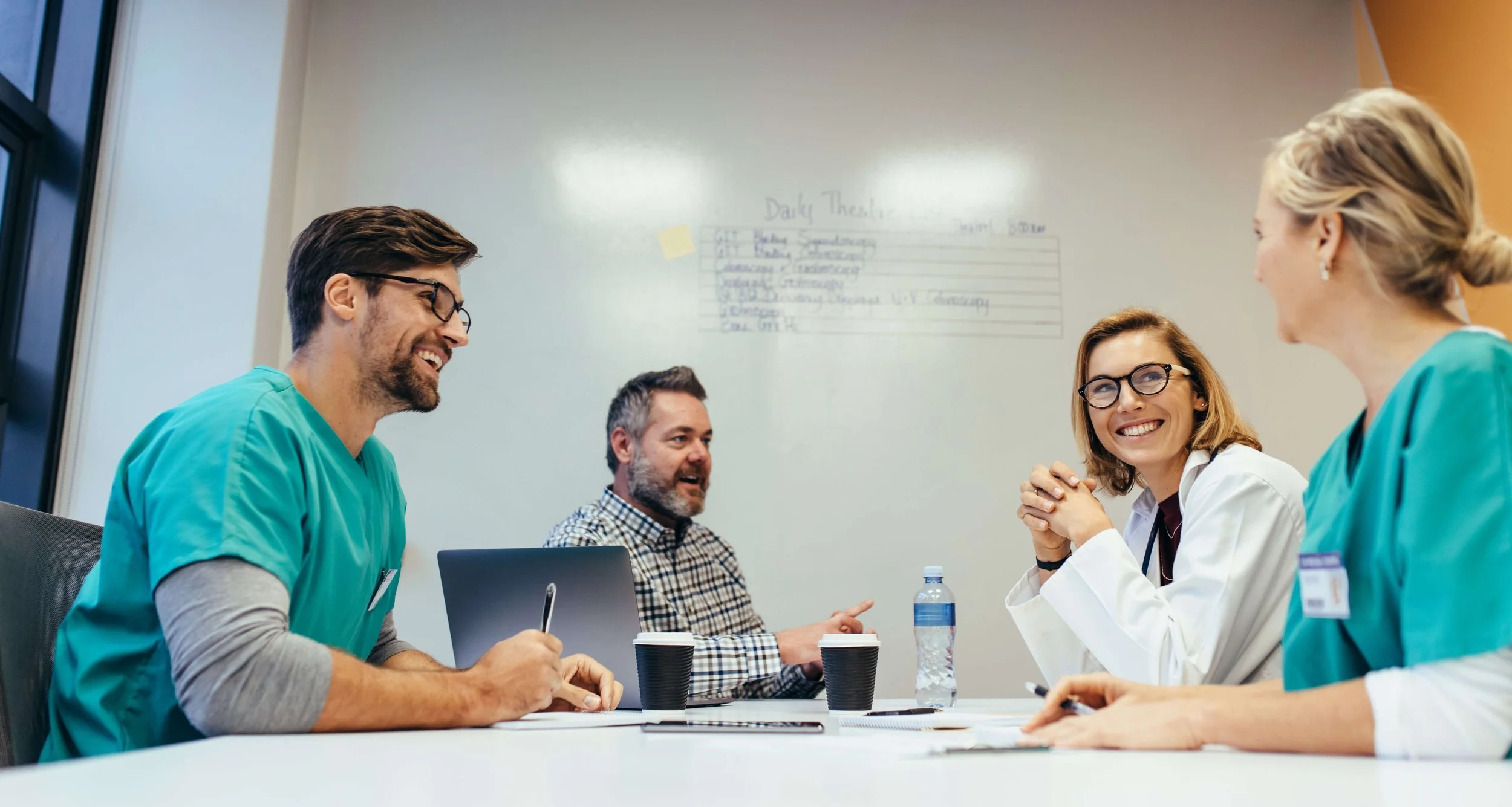 Medical professionals and a man in a meeting room, engaging in a discussion, with a whiteboard in the background.
