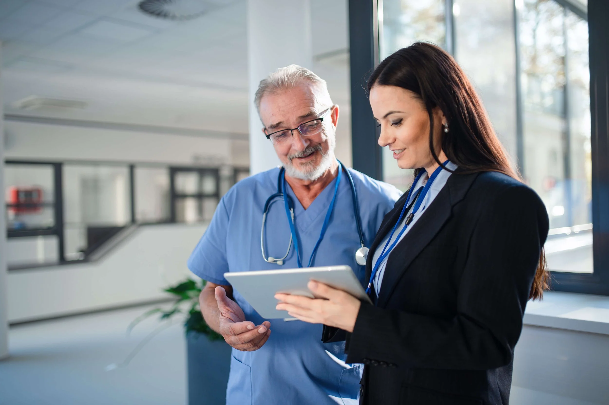 Two healthcare professionals, a senior male doctor in scrubs and a young female doctor in a suit, are looking at a tablet together in a clinic or hospital setting.