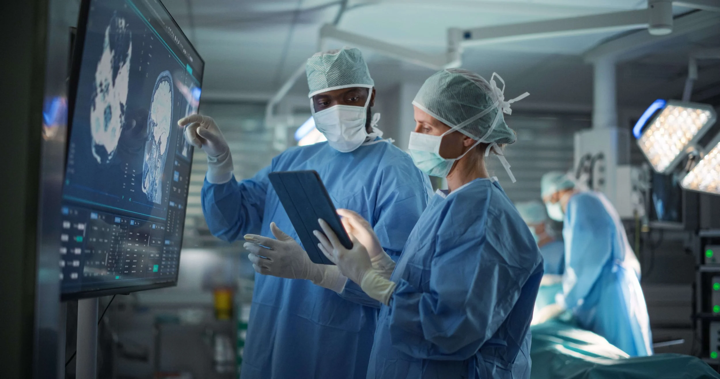 Two surgeons in scrubs, masks, and caps review a digital medical scan on a large monitor during surgery, with other medical staff and equipment in the background.