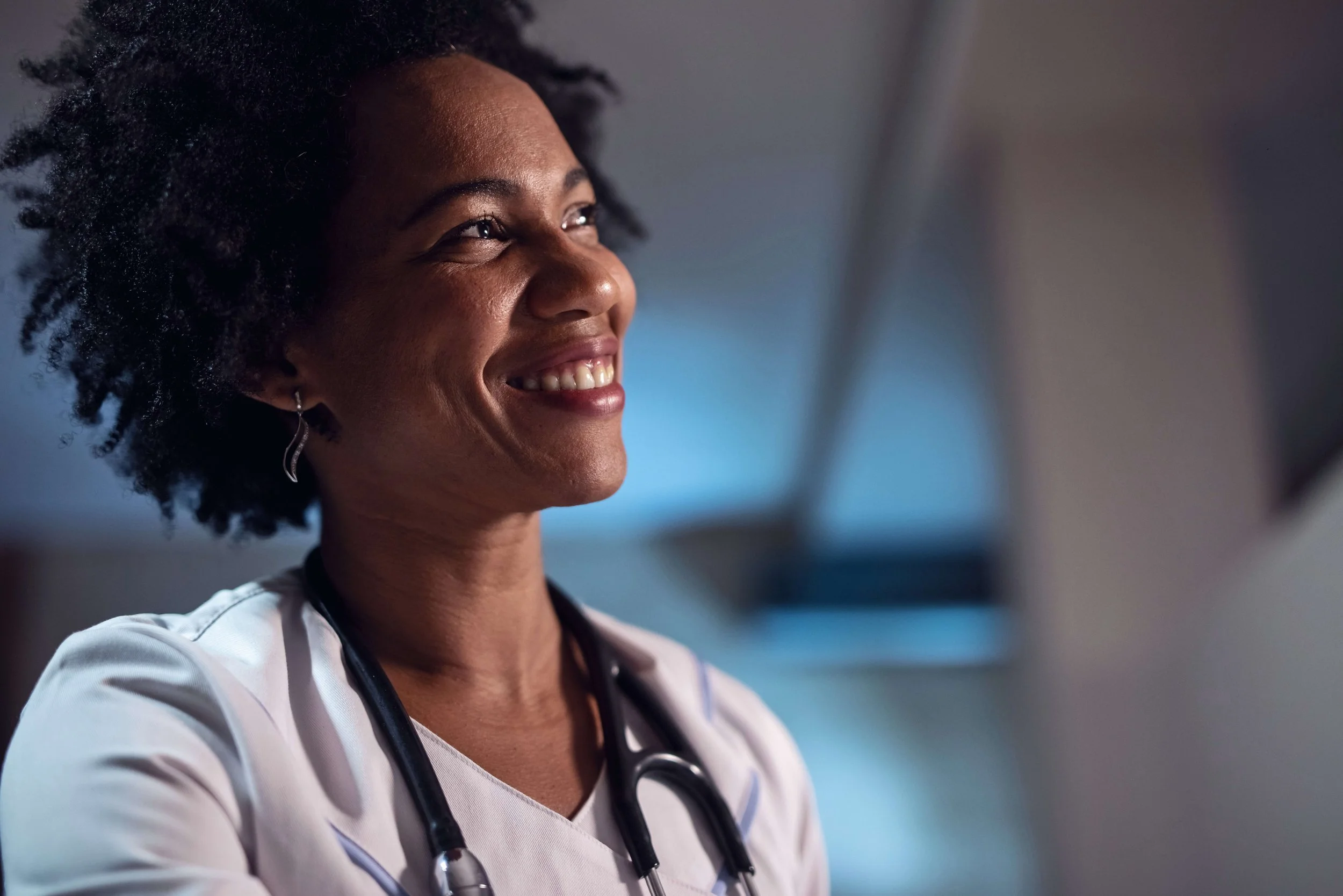 A smiling African American female nurse wearing a white uniform and stethoscope in a medical setting.