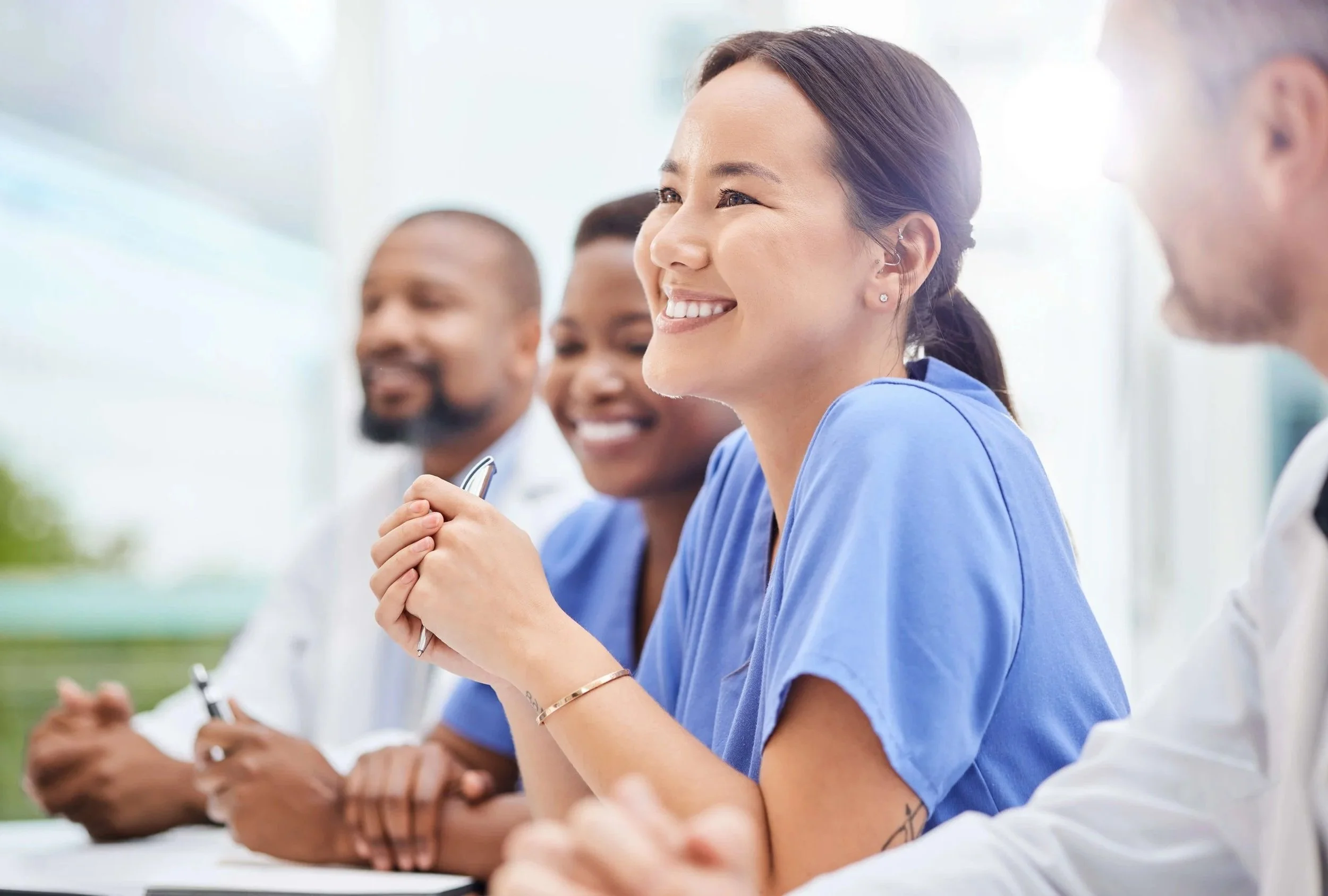 A group of diverse medical professionals in scrubs and lab coats sitting at a conference table during a meeting, smiling and engaging with each other.