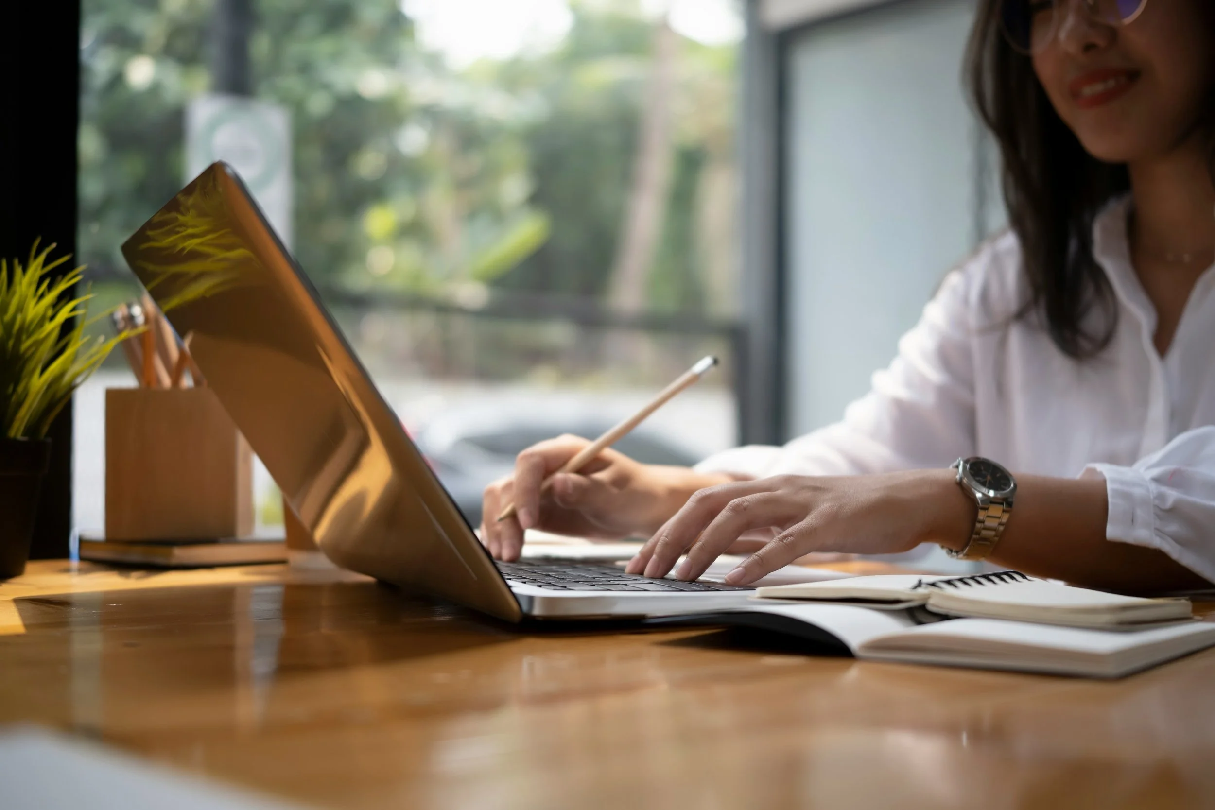 A woman working on a laptop at a wooden desk with plants and notebooks, near a large window with trees outside.