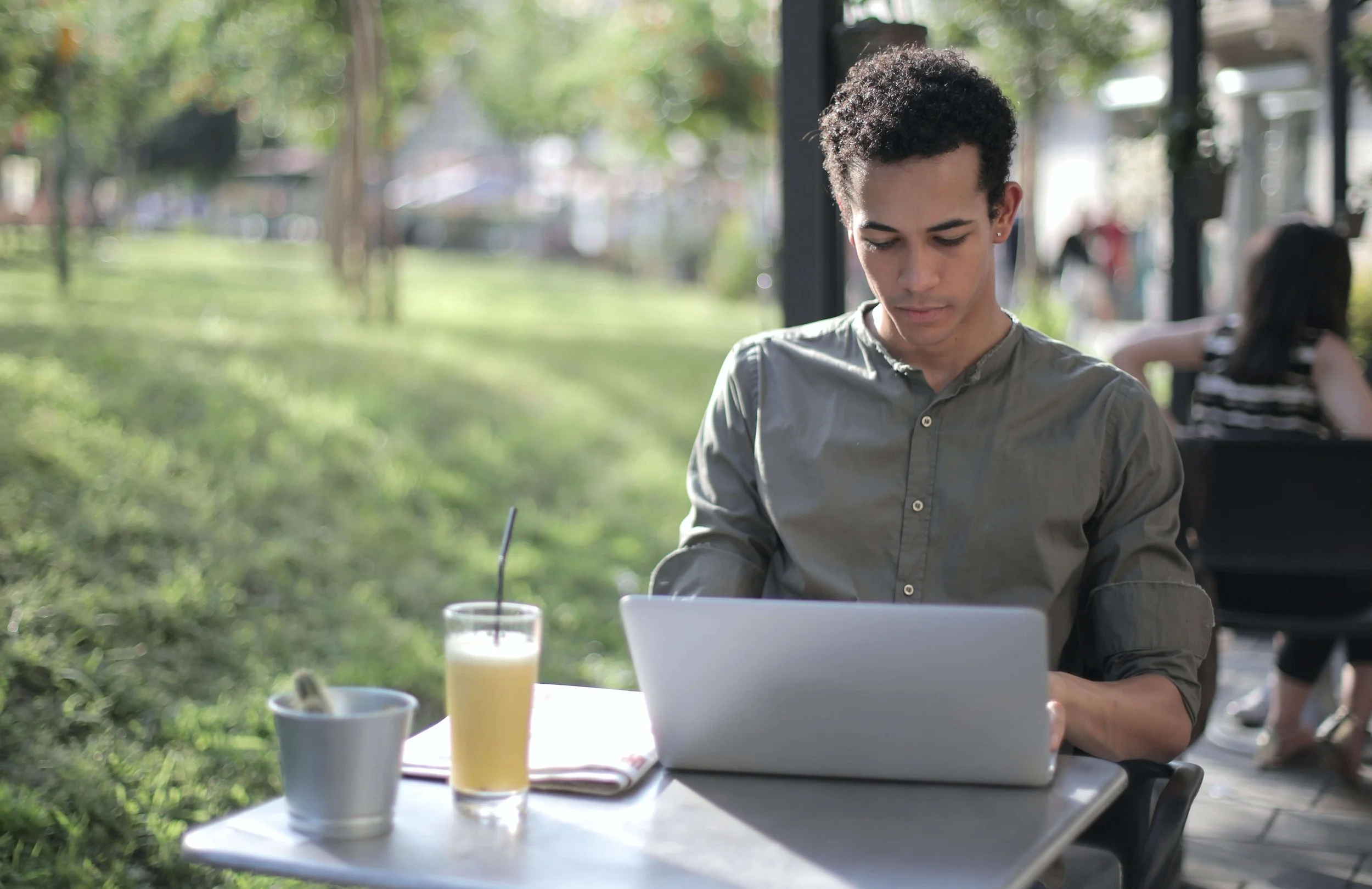 A young man working on a laptop at an outdoor cafe table with a glass of yellow juice, a white cup, and a notebook, sitting amidst green trees and other people in the background.