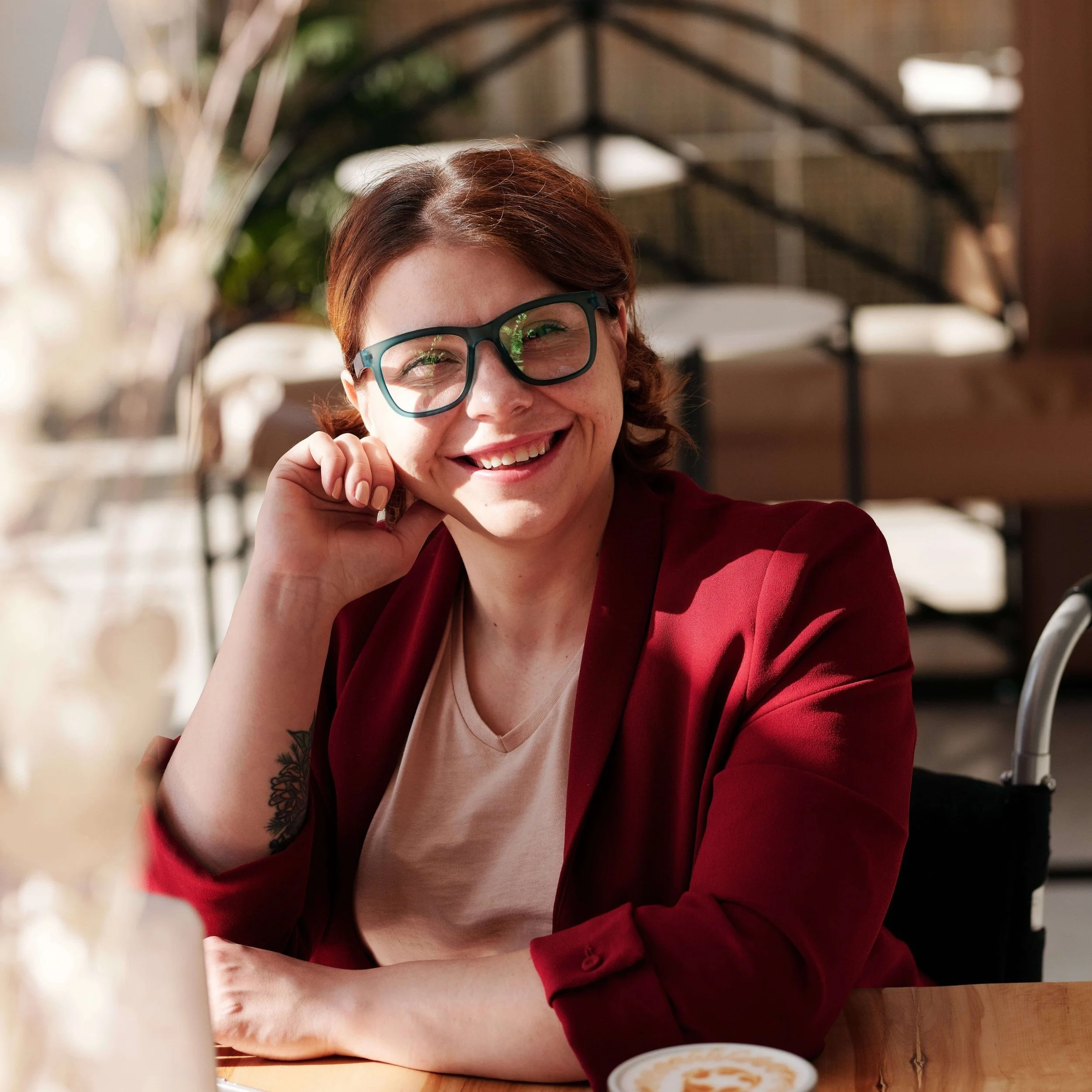 Smiling woman with red hair, glasses, and a tattoo on her wrist, sitting at a table in a well-lit cafe or restaurant.